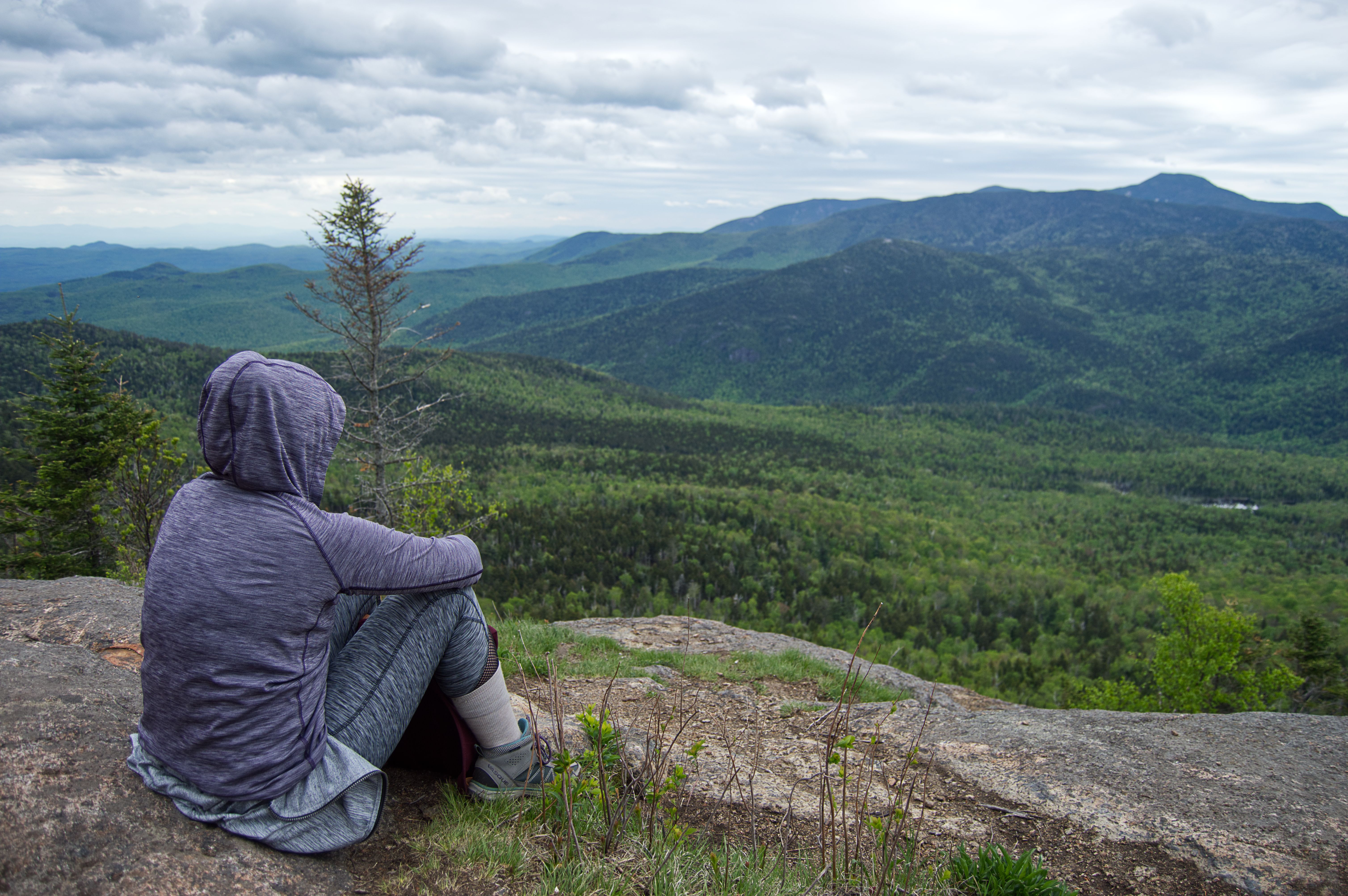 a woman in a hoodie sitting on a rock while overlooking green mountains in the distance