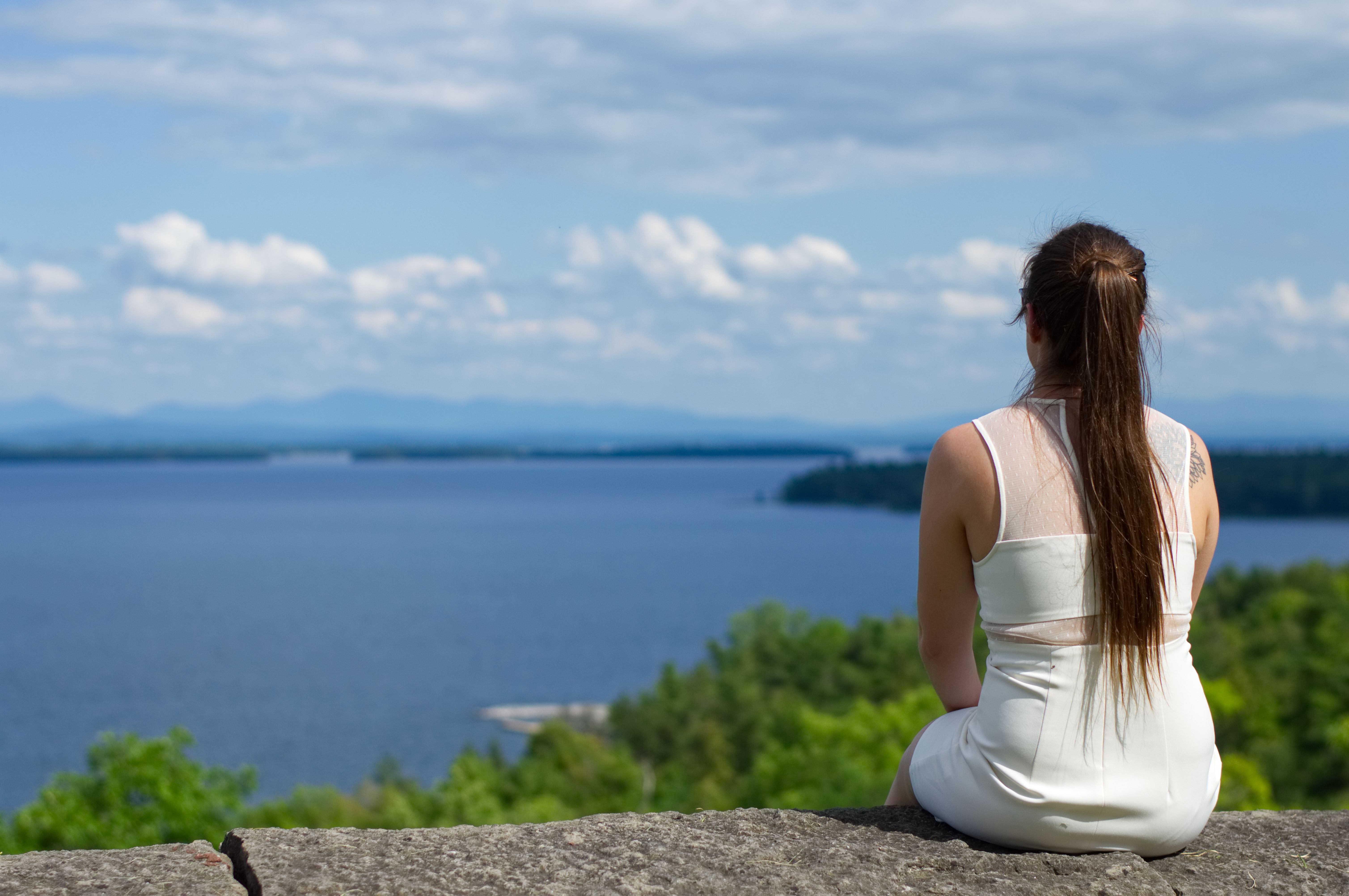 a woman in a white dress and long hair sitting on a stone while looking out over a lake