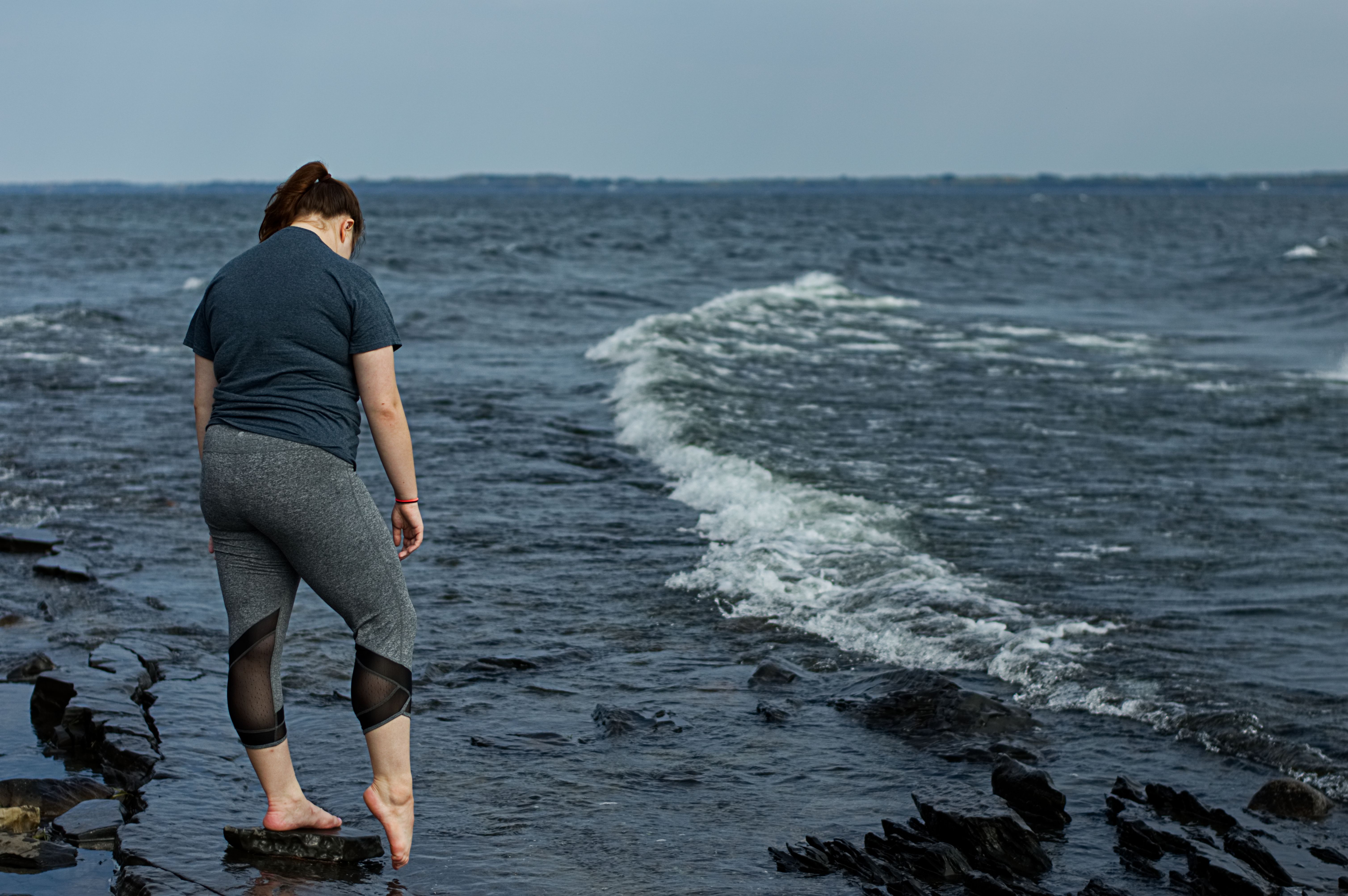 a woman dipping a single toe into water along a rocky lake shore