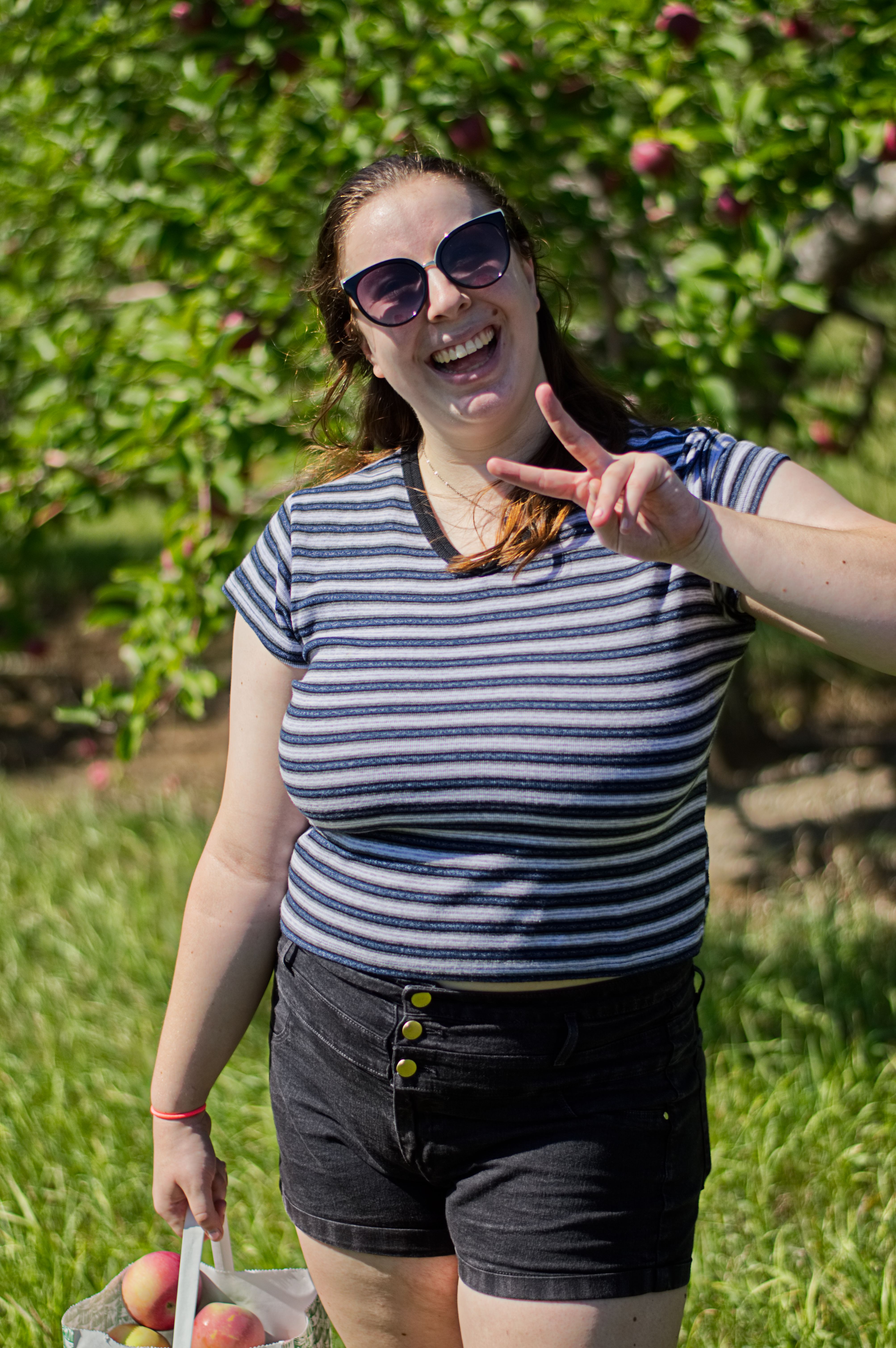 A woman in sunglasses making the peace sign with one hand, and holding a bag of apples in the other