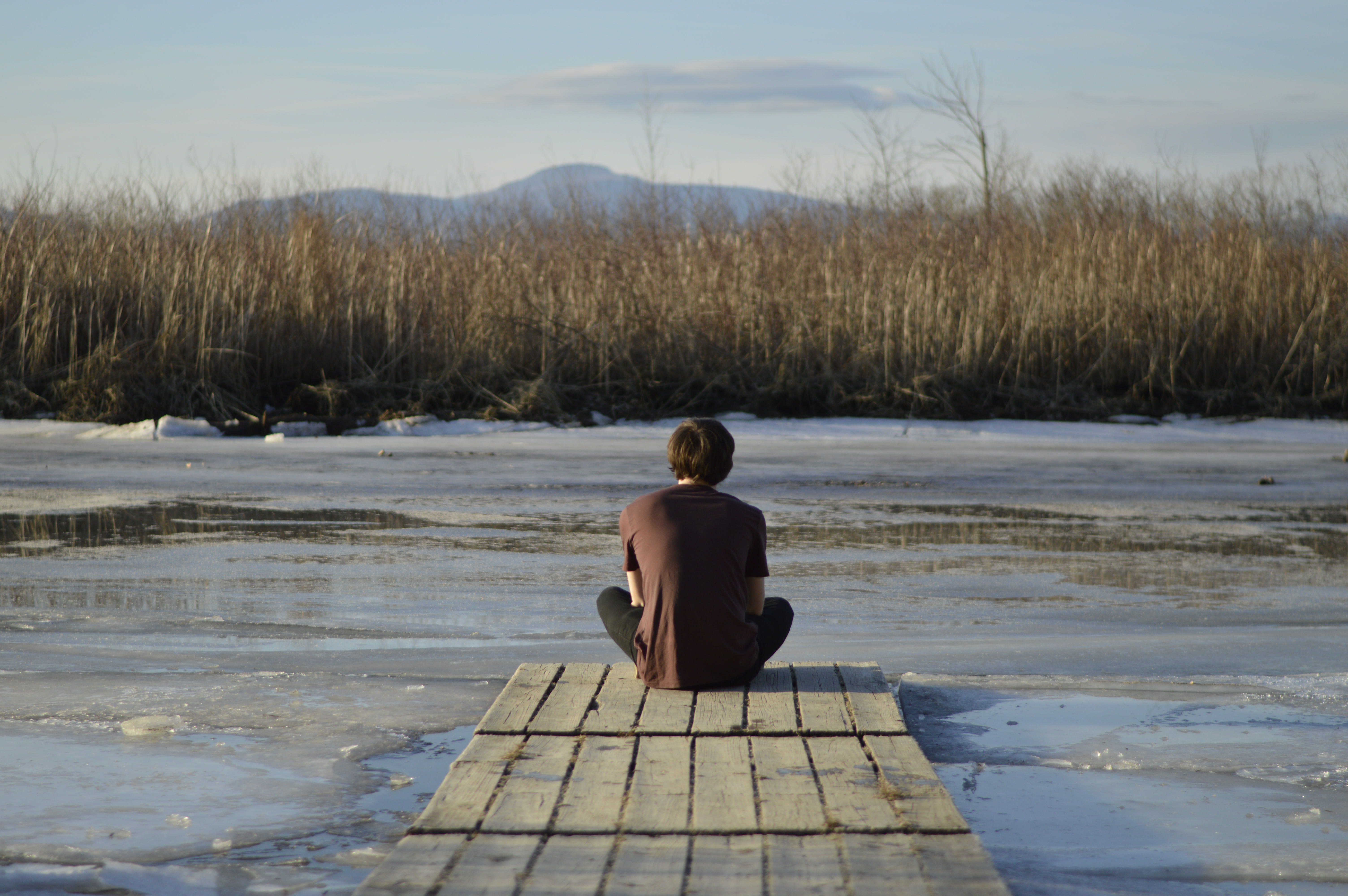 a man sitting on the end of a pier, looking out over some partially frozen water and dry tall grass