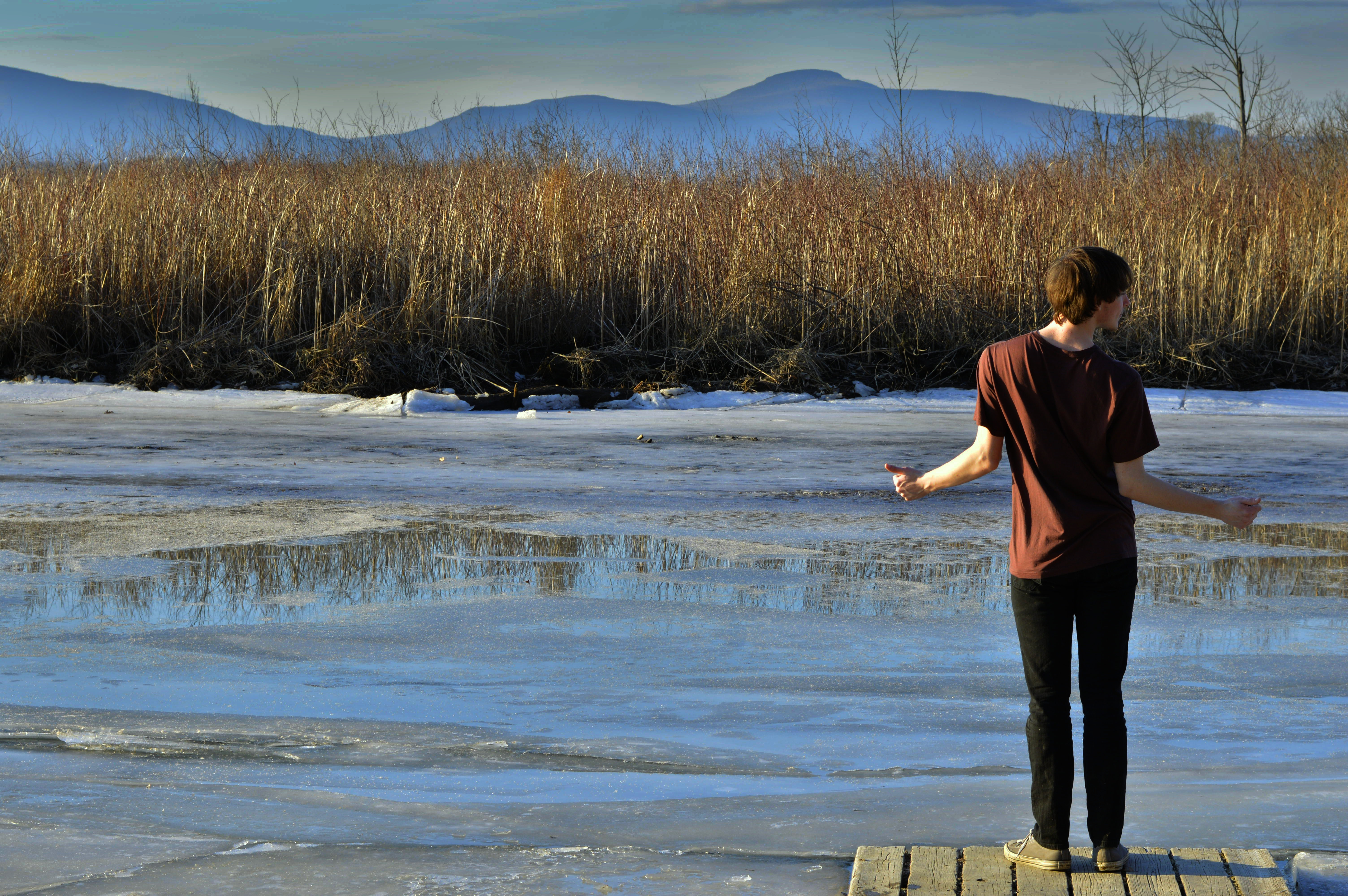 a man standing at the end of a pier looking over some partially frozen water and dry tall grass