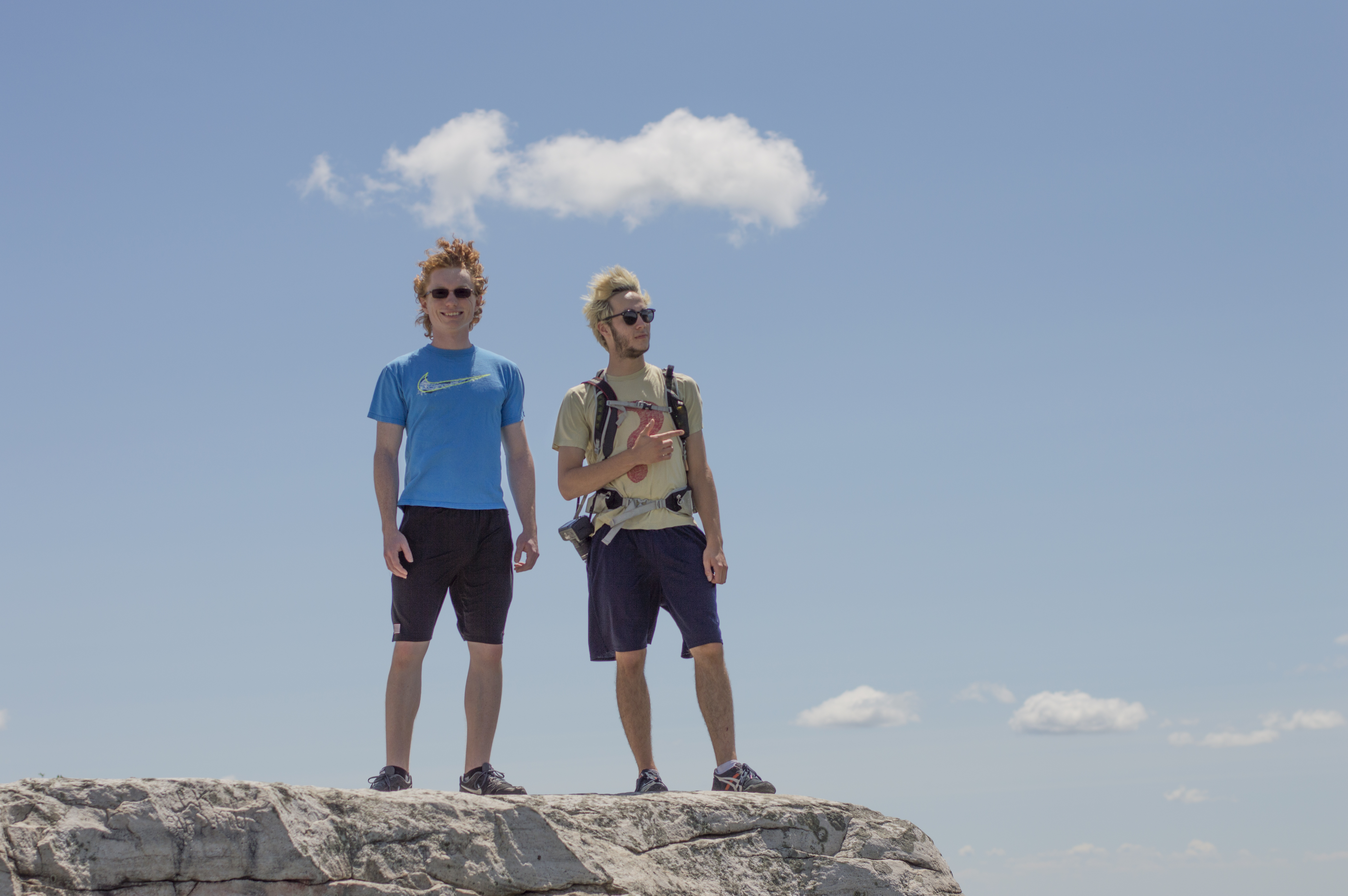 Two men standing atop a rock with nothing but the sky behind them. one looks towards the camera, the other points off to the right where they are looking