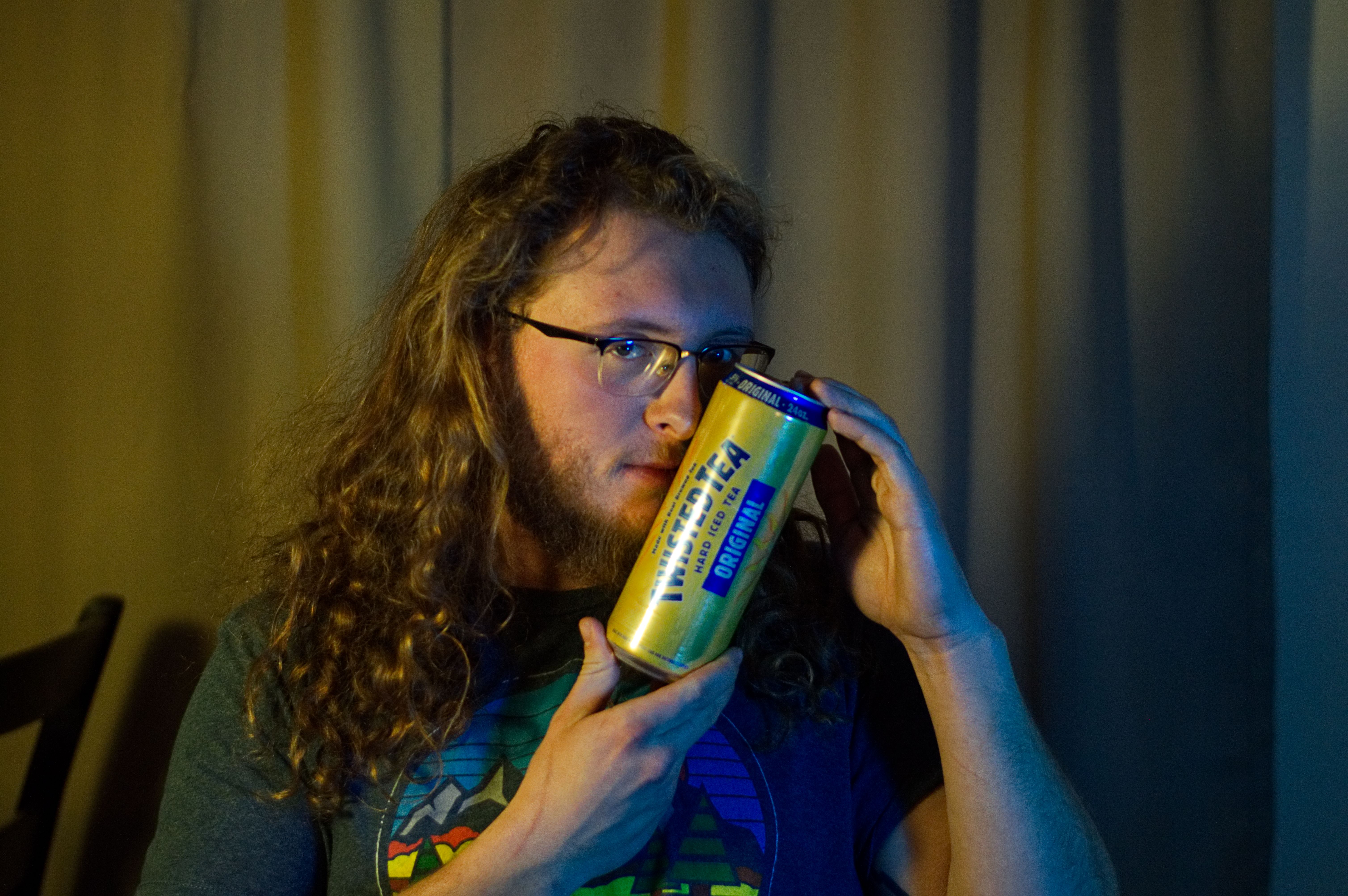 A man with long hair and a beard holding a Twisted Tea Hard Iced Tea can to his face while staring at the camera