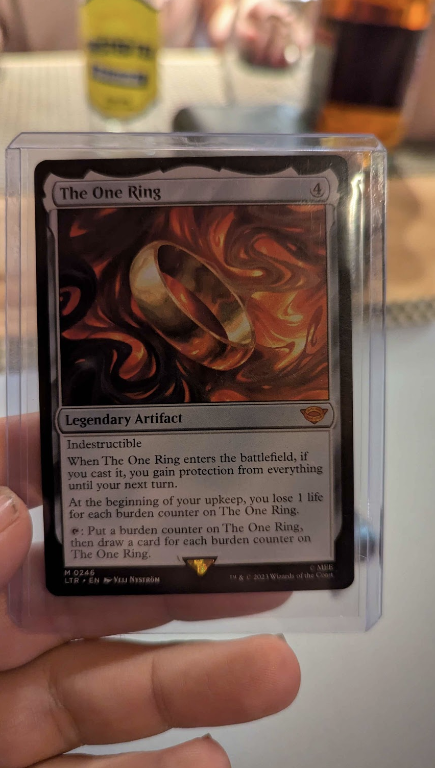 a photo of the Magic the Gathering Card 'The One Ring'. It is a legendary artifact that costs 4 colorless mana. It is indestructible, and has the following effects. 1 - When The One Ring enters the battlefield, if you cast it, you gain portection from everything until your next turn. 2- At the beginning of your upkeep, you lose 1  life for each burden counter on The One Ring. 3- Tap The One Ring, and put a burden counter on it. Draw 1 card for each burden counter on The One Ring.