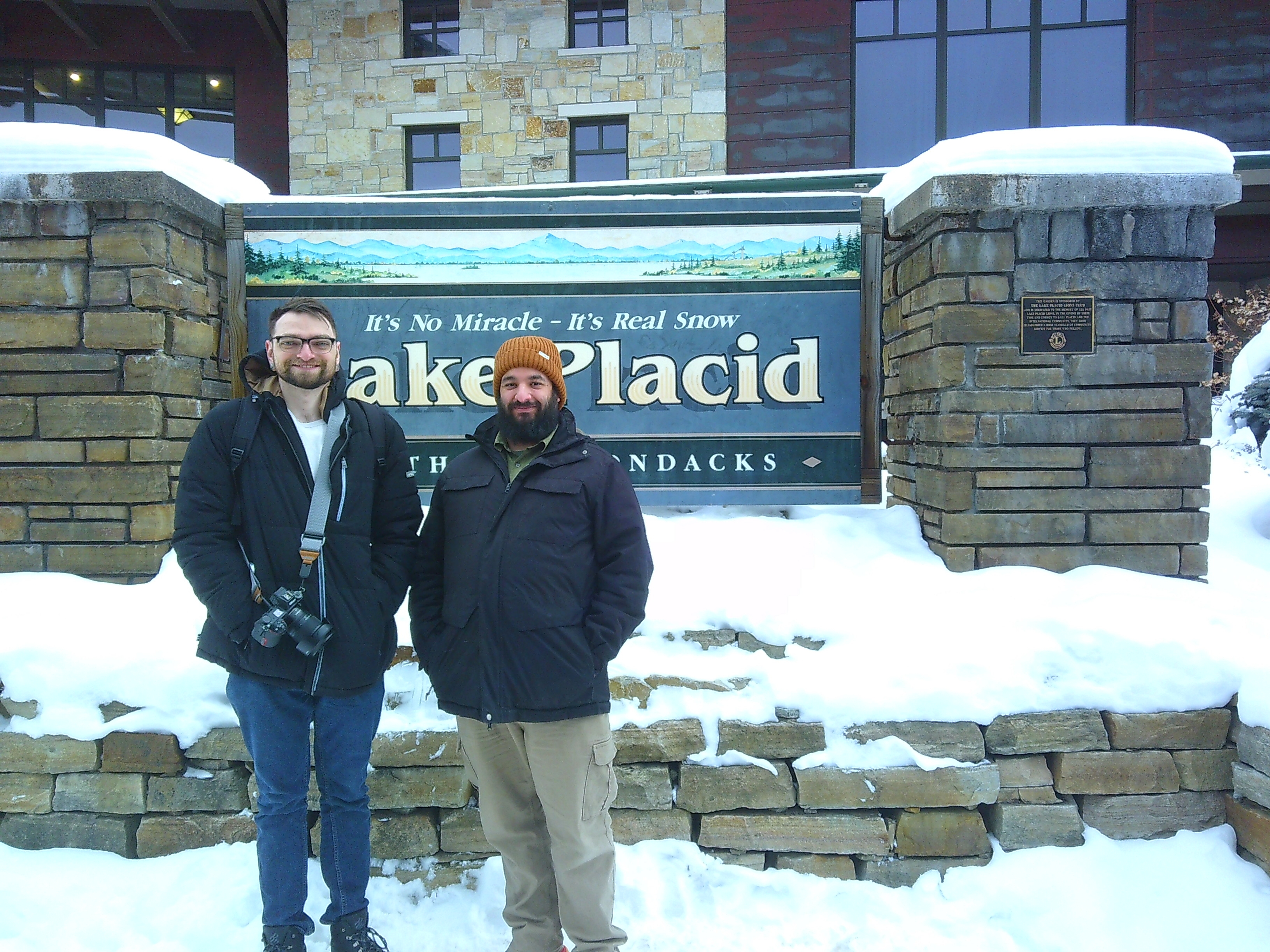 photo of me and Tanner standing in front of the Lake Placid sign. He has his camera on a strap going diagonal from his neck under his right arm