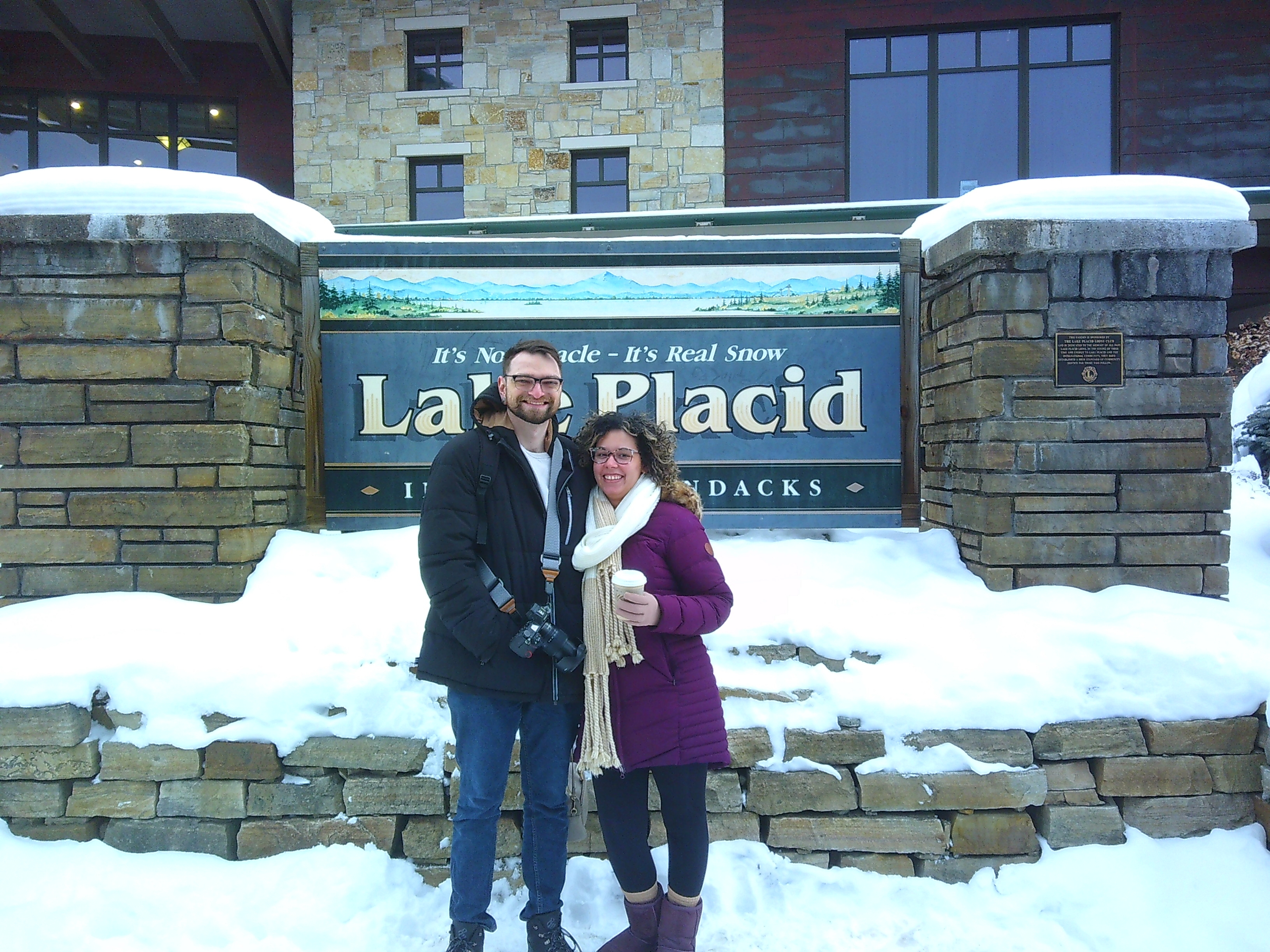 photo of Tanner and Ali in front of the Lake Placid sign. They're both smiling, Tanner has his camera around his neck and under his arm, Ali is wearing a purple coat with purple boots while holding a coffee