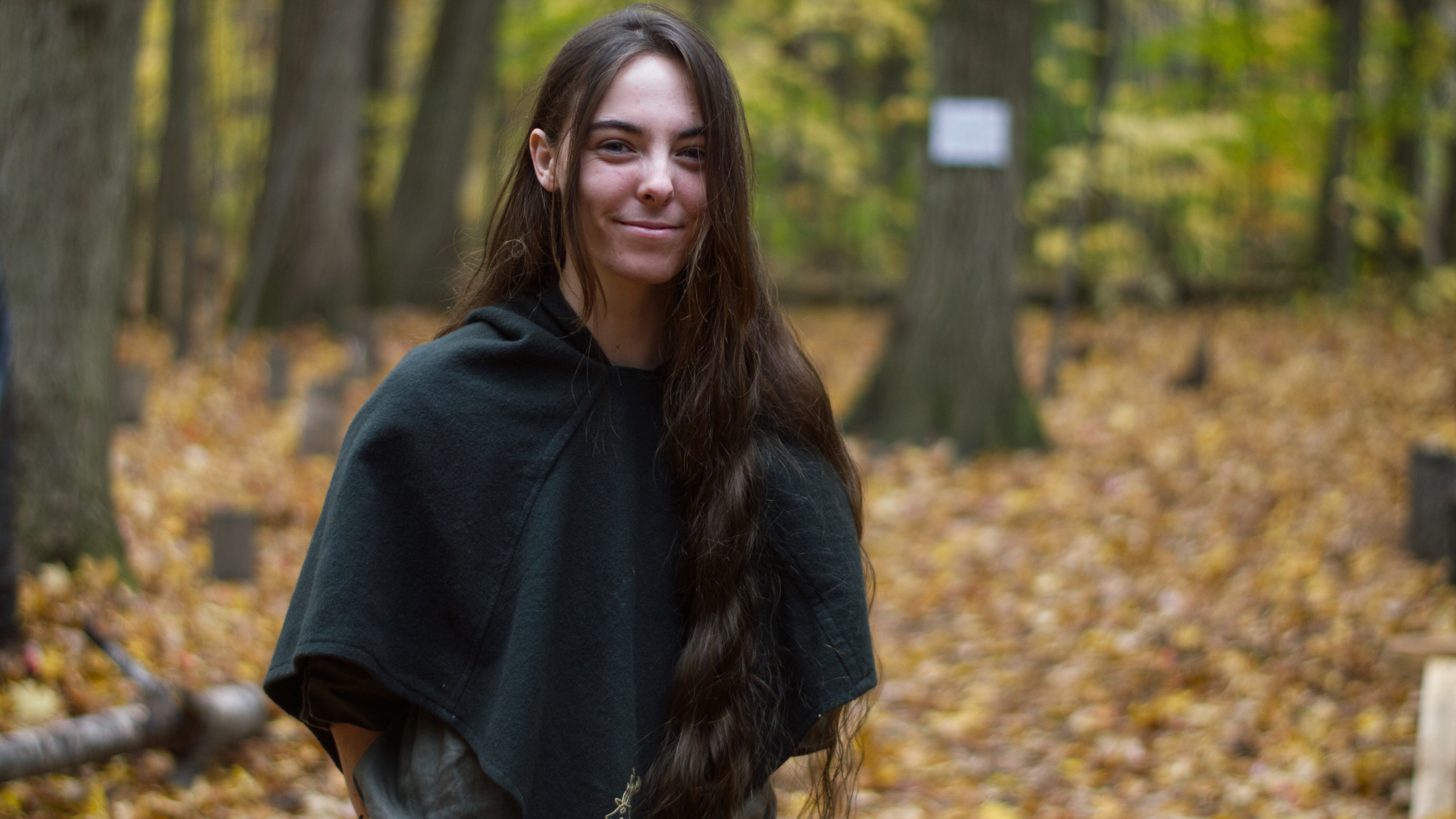 image of a person in the woods smiling. They have long hair, and are dressed like a medeival ranger.