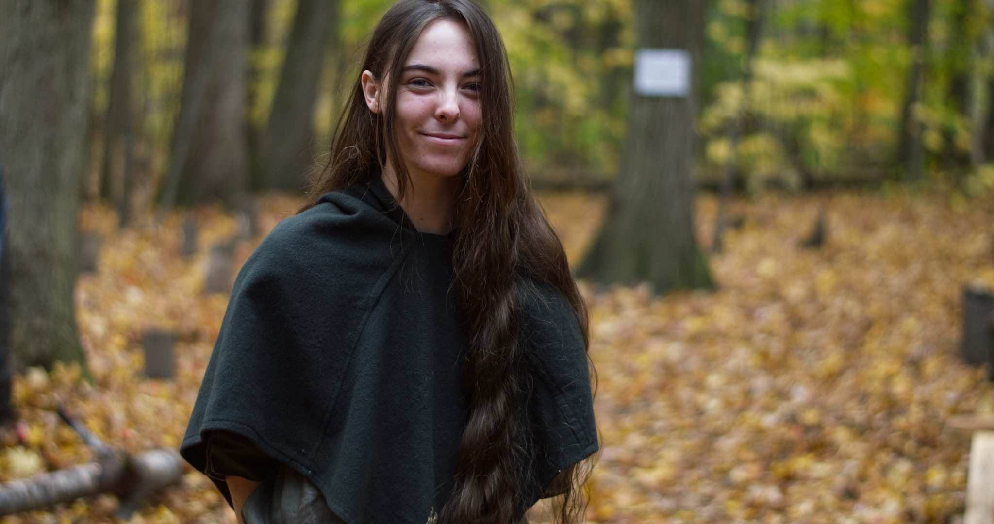 image of a person in the woods smiling. They have long hair, and are dressed like a medeival ranger.