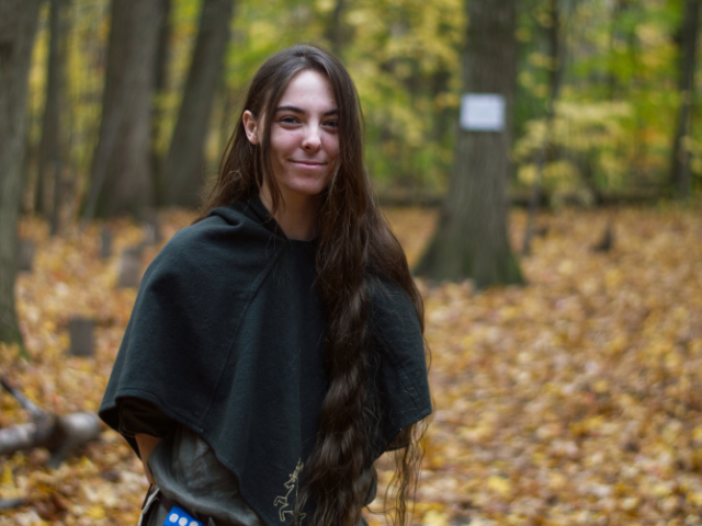 image of a person in the woods smiling. They have long hair, and are dressed like a medeival ranger.