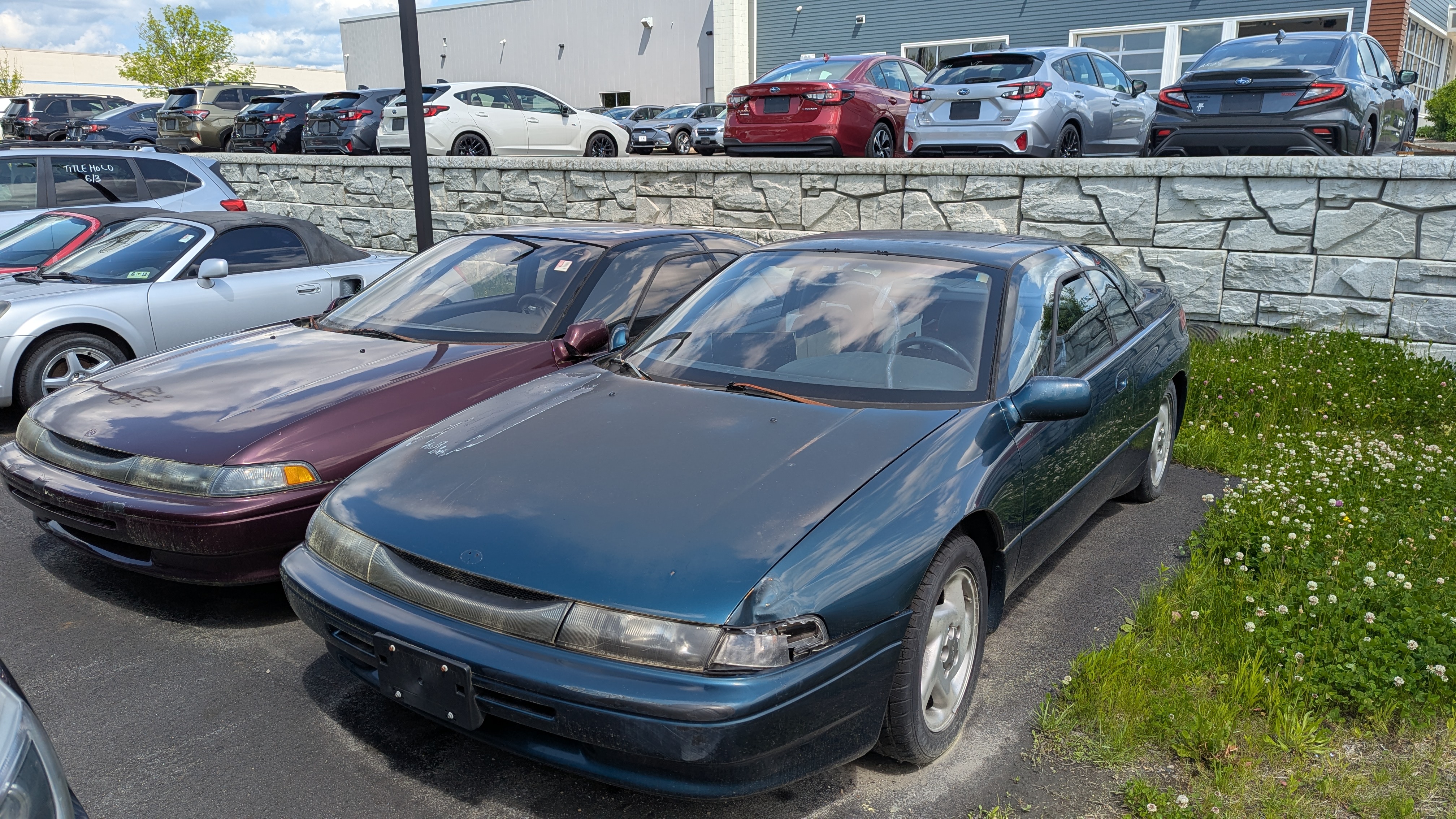 Photograph of two Subaru SVXs from the front, one green ish one purple ish. They're slightly damaged, and sitting on a car lot with a bunch of other cars