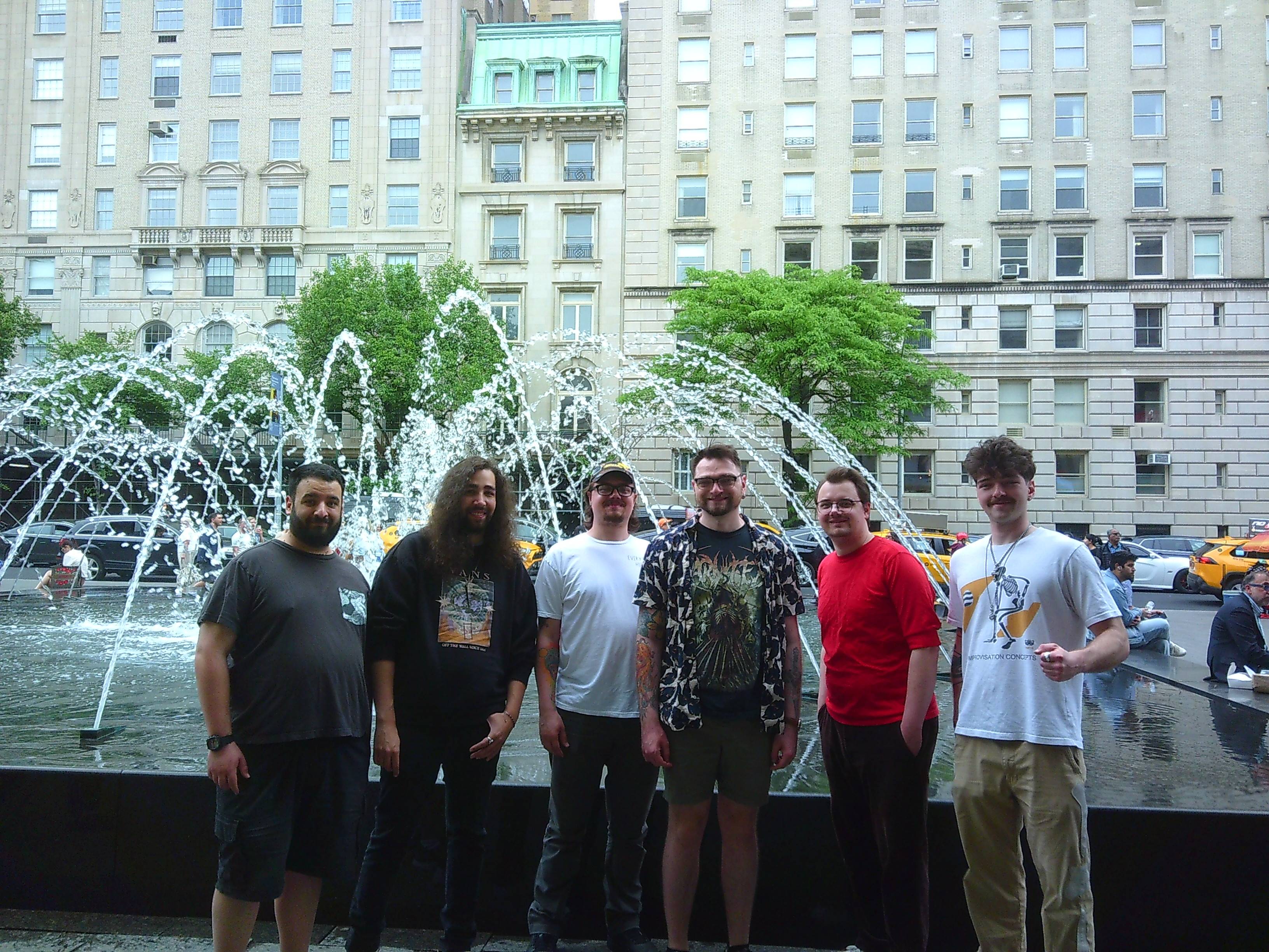 From left to right, Steven, Ben, Aiden, Tanner, Dylan, and Skyler standing in front of a fountain in NYC. A white building can be seen behind them