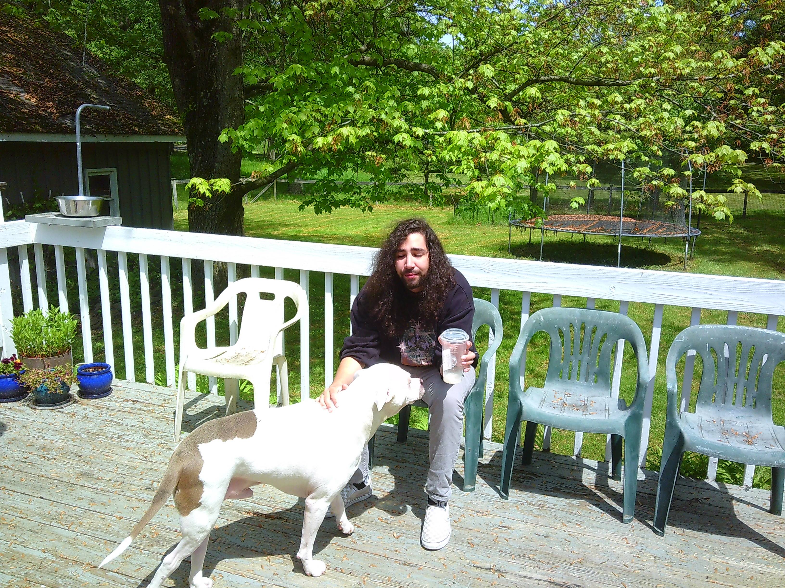 a man with long hair in long pants and a long sweatshirt sitting on a back porch petting a pitbull looking dog. The dog is mostly white with some brown near it's rear. There is a field behind where the man is sitting, with a trampoline in the background.