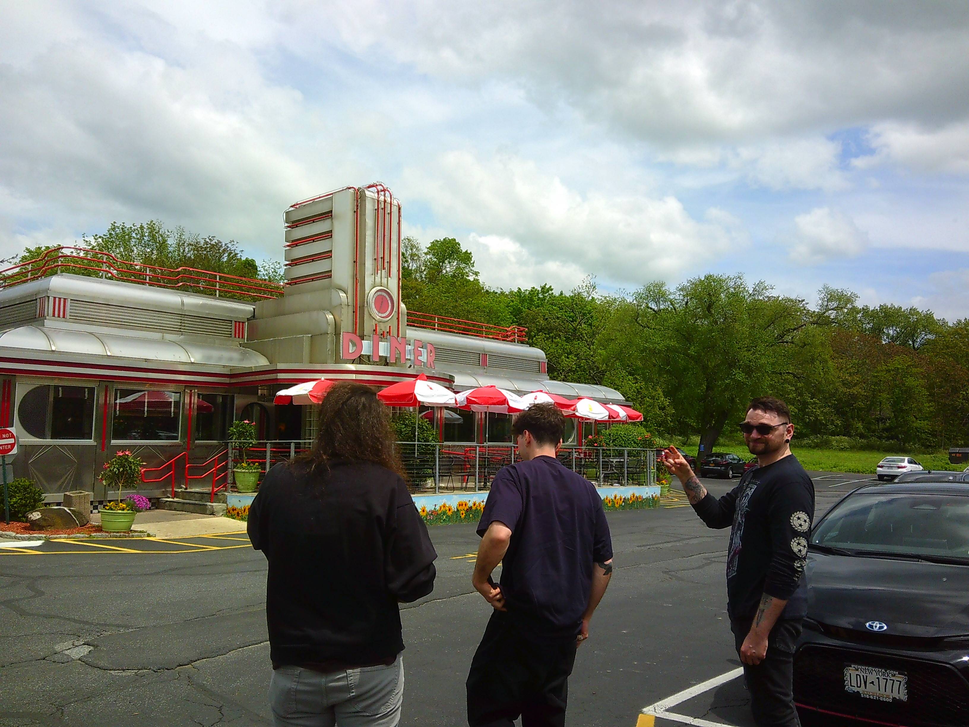 Three men, two facing away from camera another facing towards the camera, heading to a retro looking building that says Diner. The building is largely silver and Red