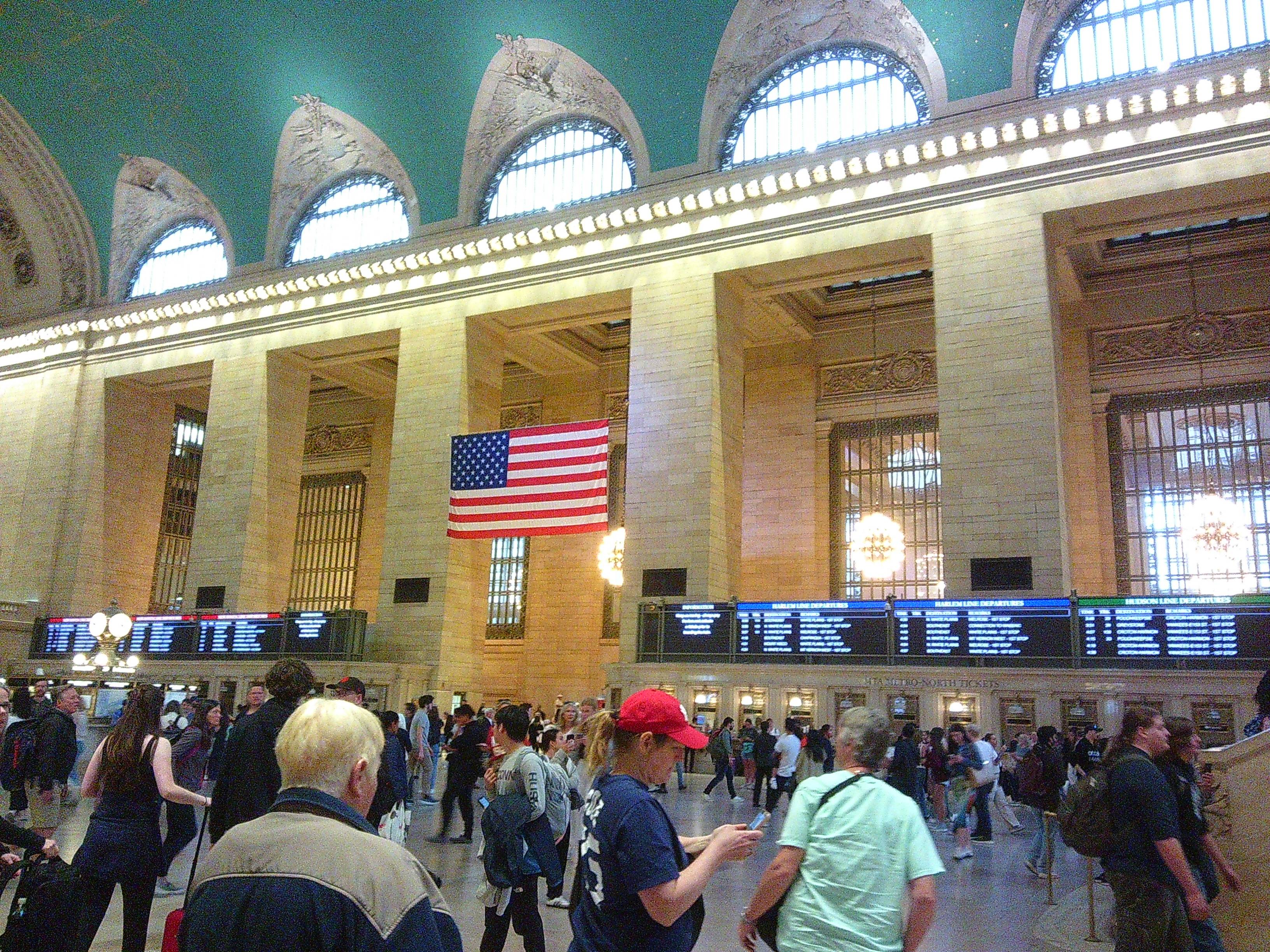 Picture of a lot of people inside of Grand Central. An American flag can be seen in the distance.