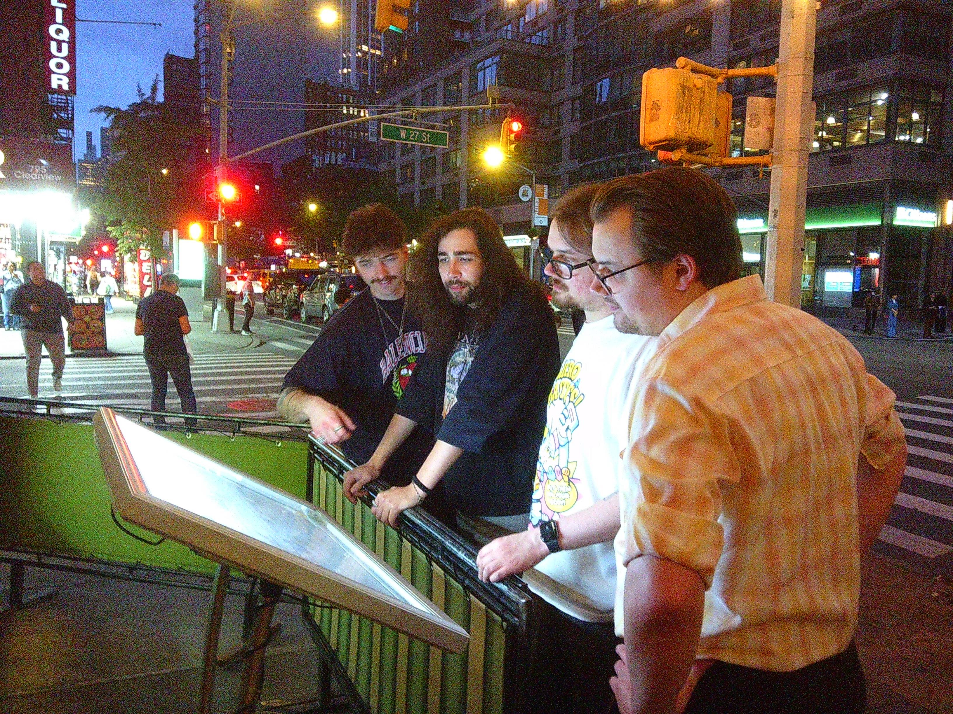 4 men (Skyler, Ben, Aiden, Dylan from left to right) looking at an illuminated menu outside of a restaurant in New York City. It is night time, and they are near an intersection.