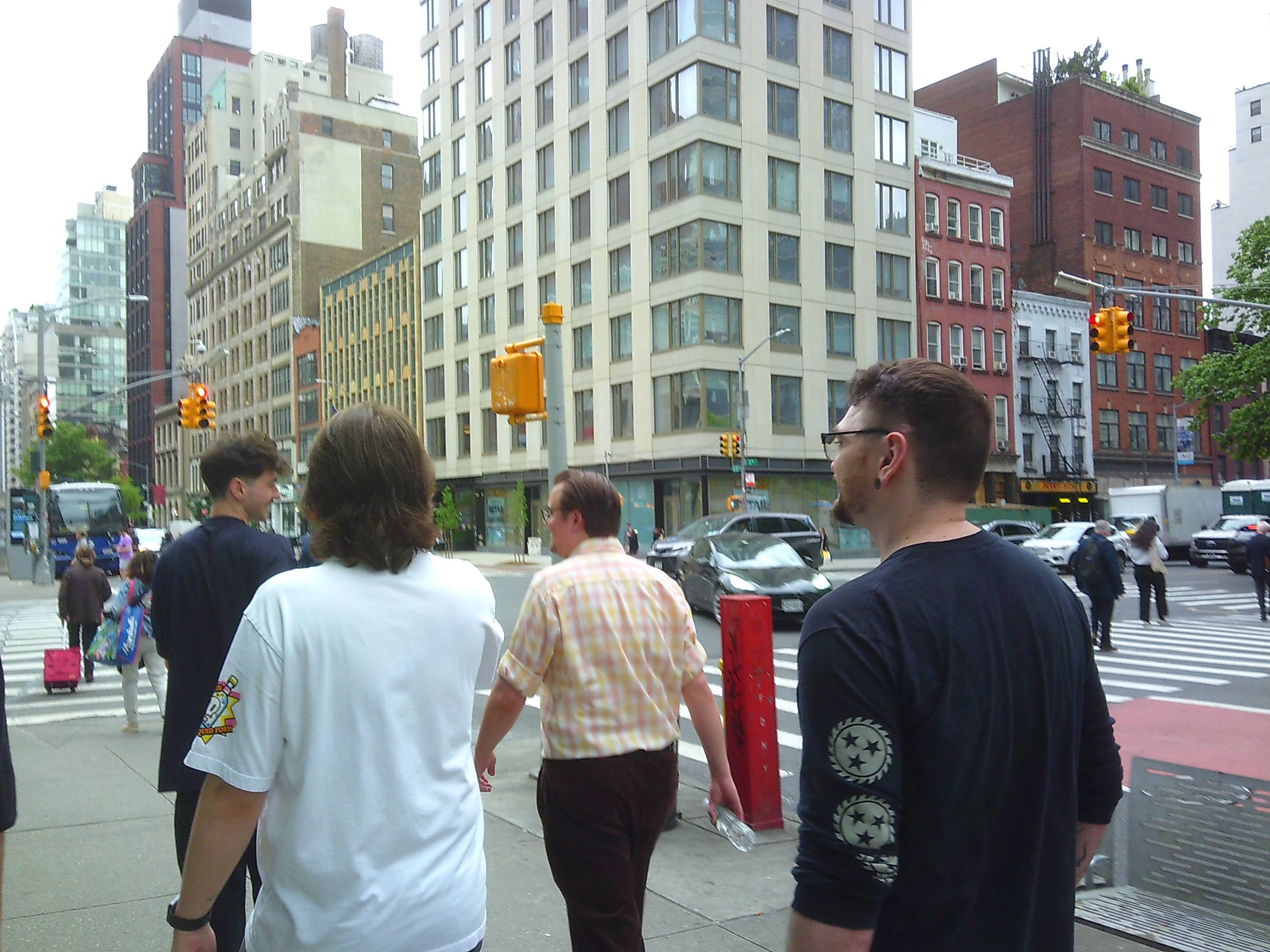 A photo from behind 4 men walking the streets of New York City. They're near an intersection, and they can be seen talking amonst each other