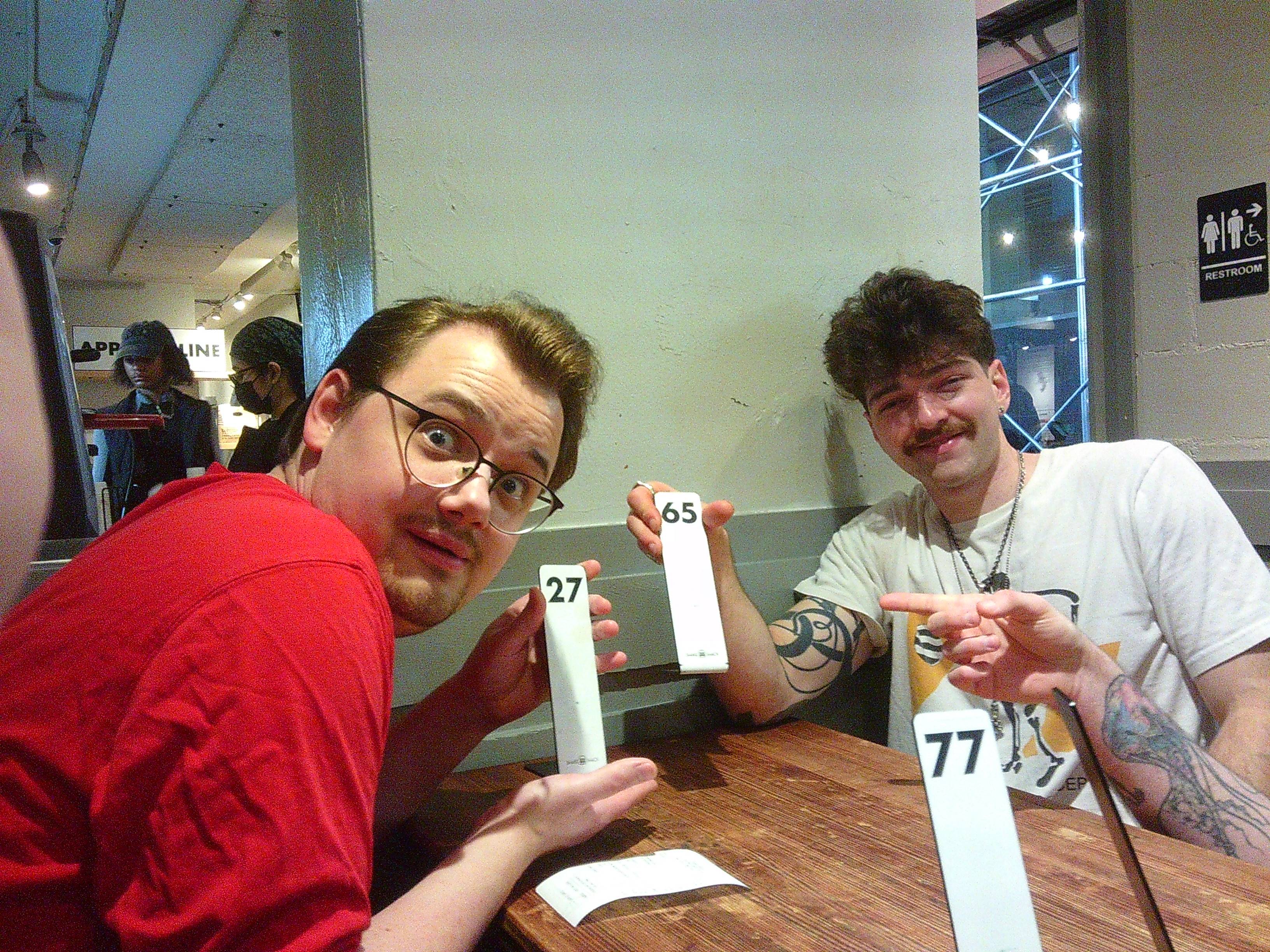 A photo from inside a restaurant, with Dylan holding a number 27 order stand thing. Skyler is holding one that is labeled 65, and someone is holding one labeled 77