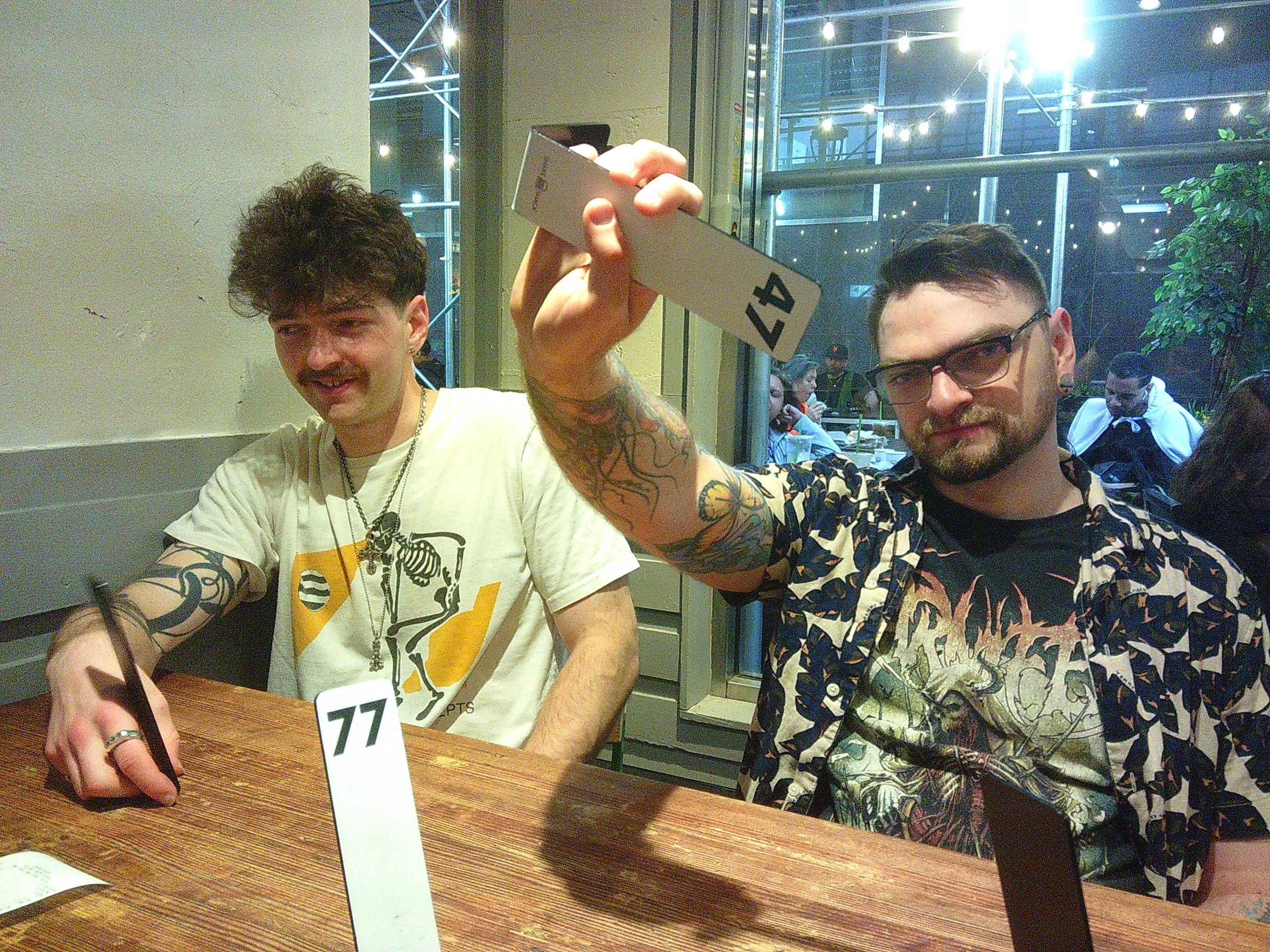 A photo from inside a restaurant, with Skyler looking off camera and Tanner holding a number 47 order stand thing. People can be seen behind Tanner in another area