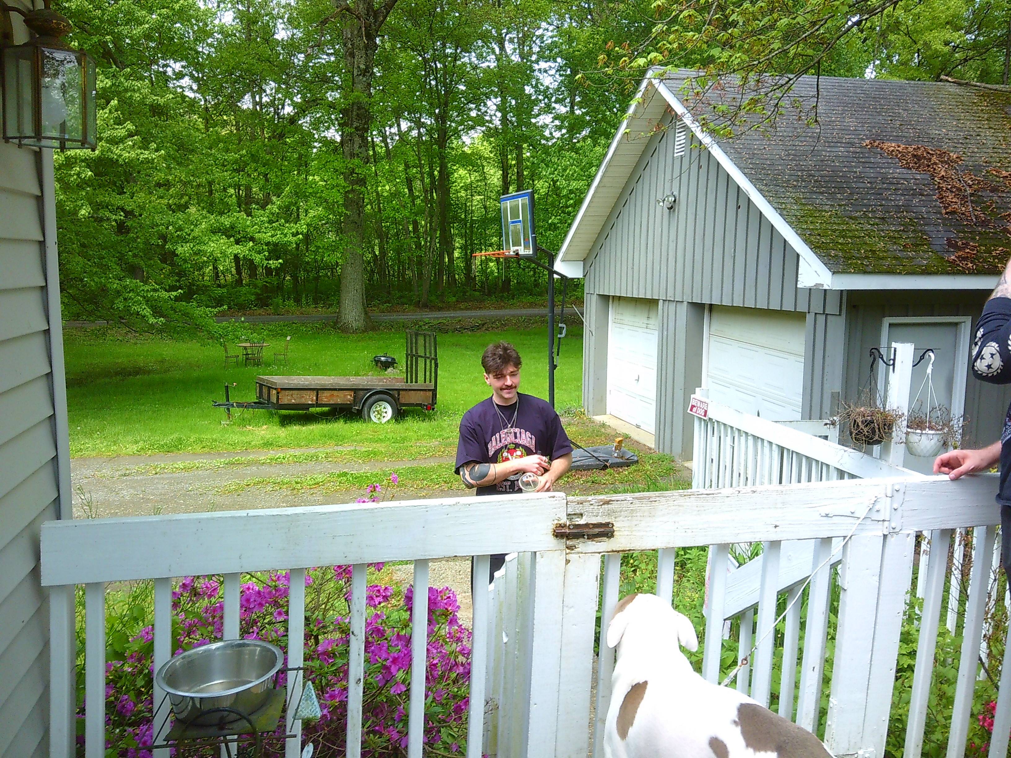 a man in a tshirt wearing some long necklaces approaching a locked fence gate to get onto a back porch, with a pitbull looking dog waiting to greet him at the gate. The dog is mostly white with some brown spots