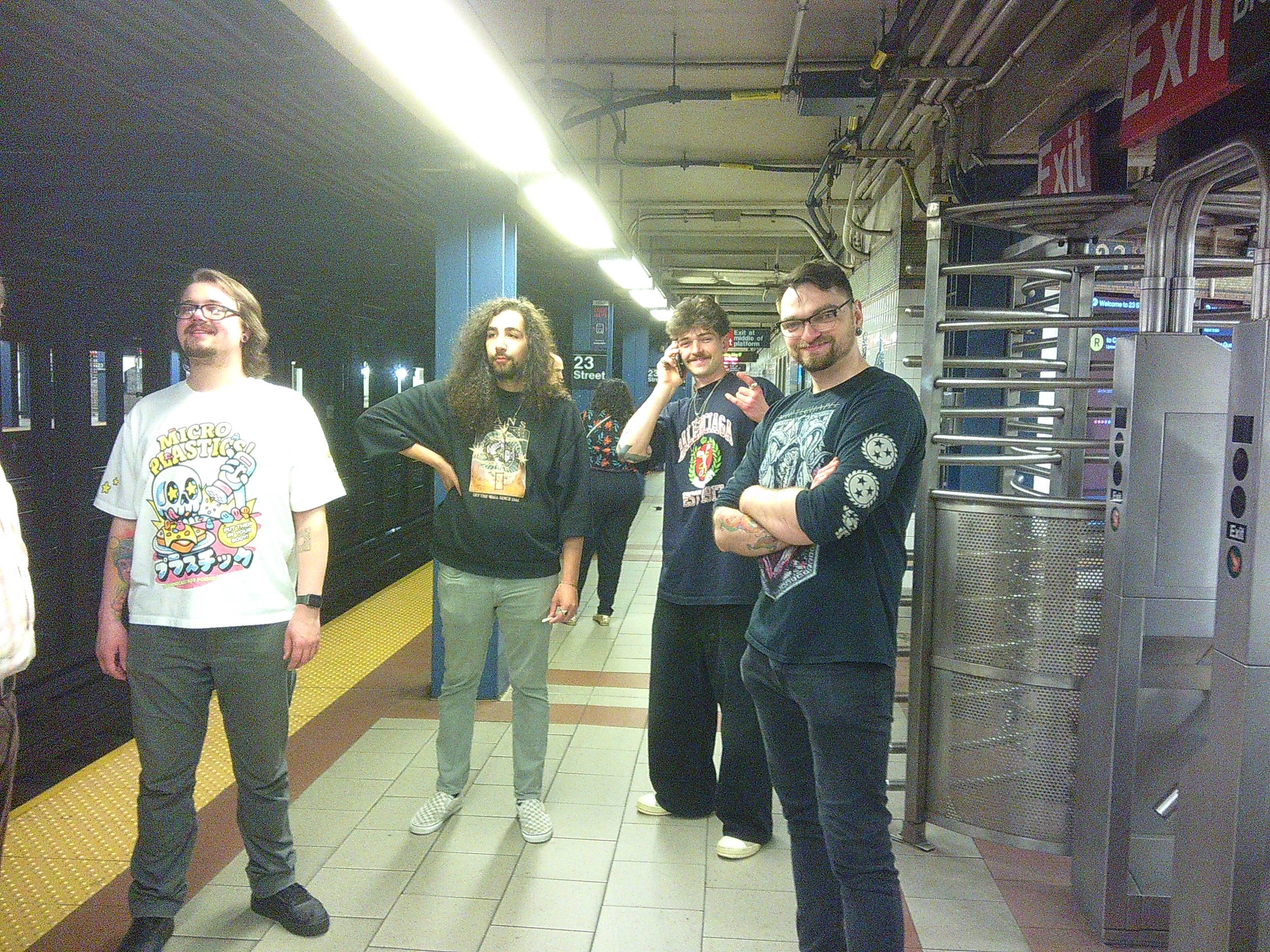 4 men (Aiden, Ben, Skyler, Tanner from left to right) waiting at a subway stop. Skykler is on his phone