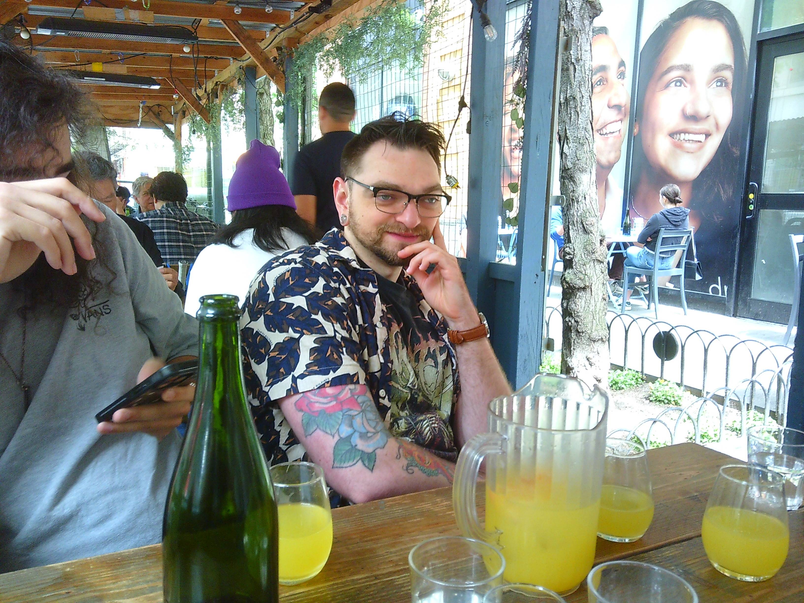 A shot of two men (Ben and Tanner) at a table outside. The table has a couple of glasses of a bright yellow drink. In the background you can see more people at other tables, as well as advertisements on the sides of buildings