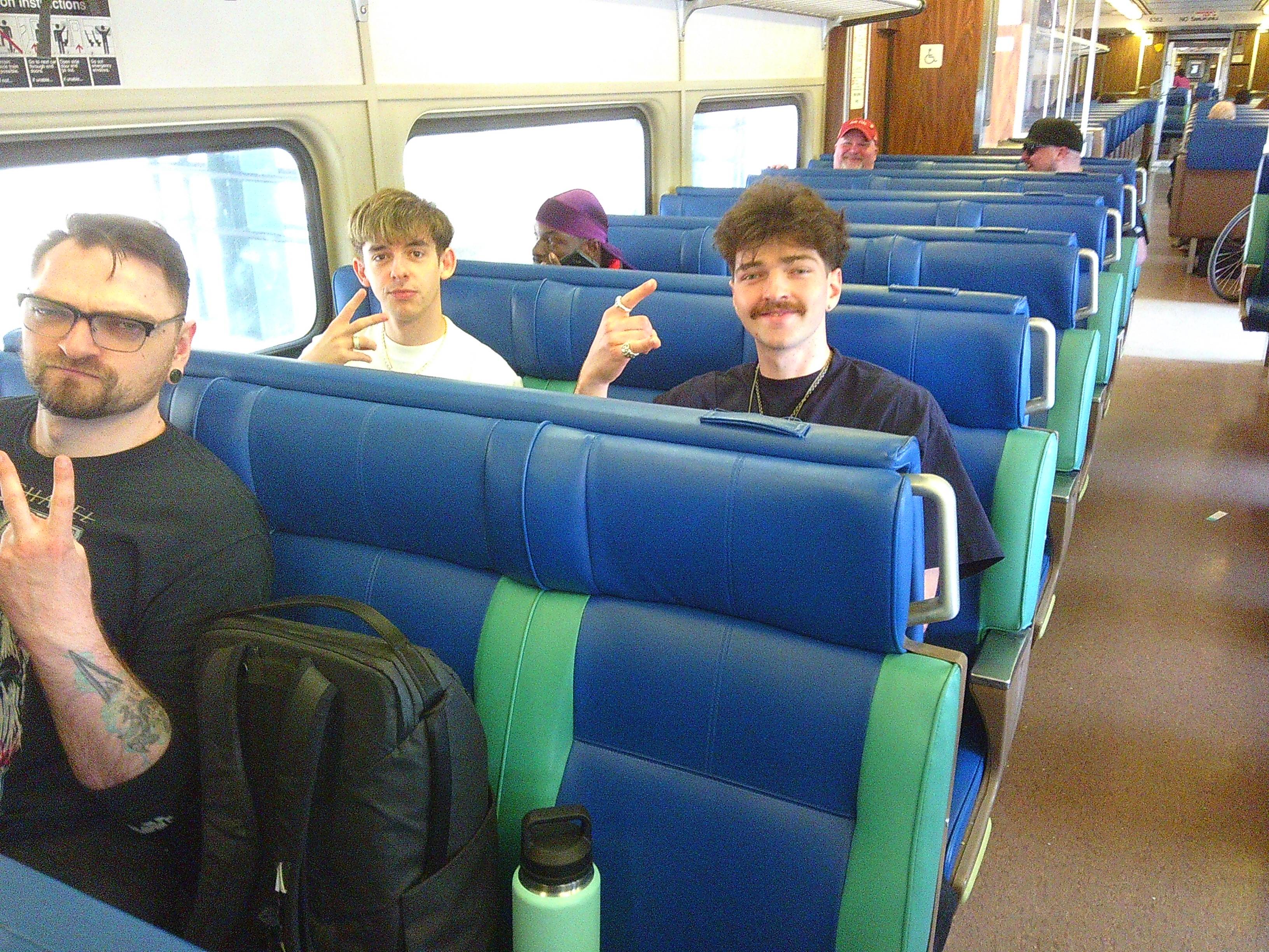 Picture looking down a train aisle. There are three men visibile in the foreground. Tanner the Groom is in the left of the photo in the front seat, with Skyler and his friend in the row behind him.