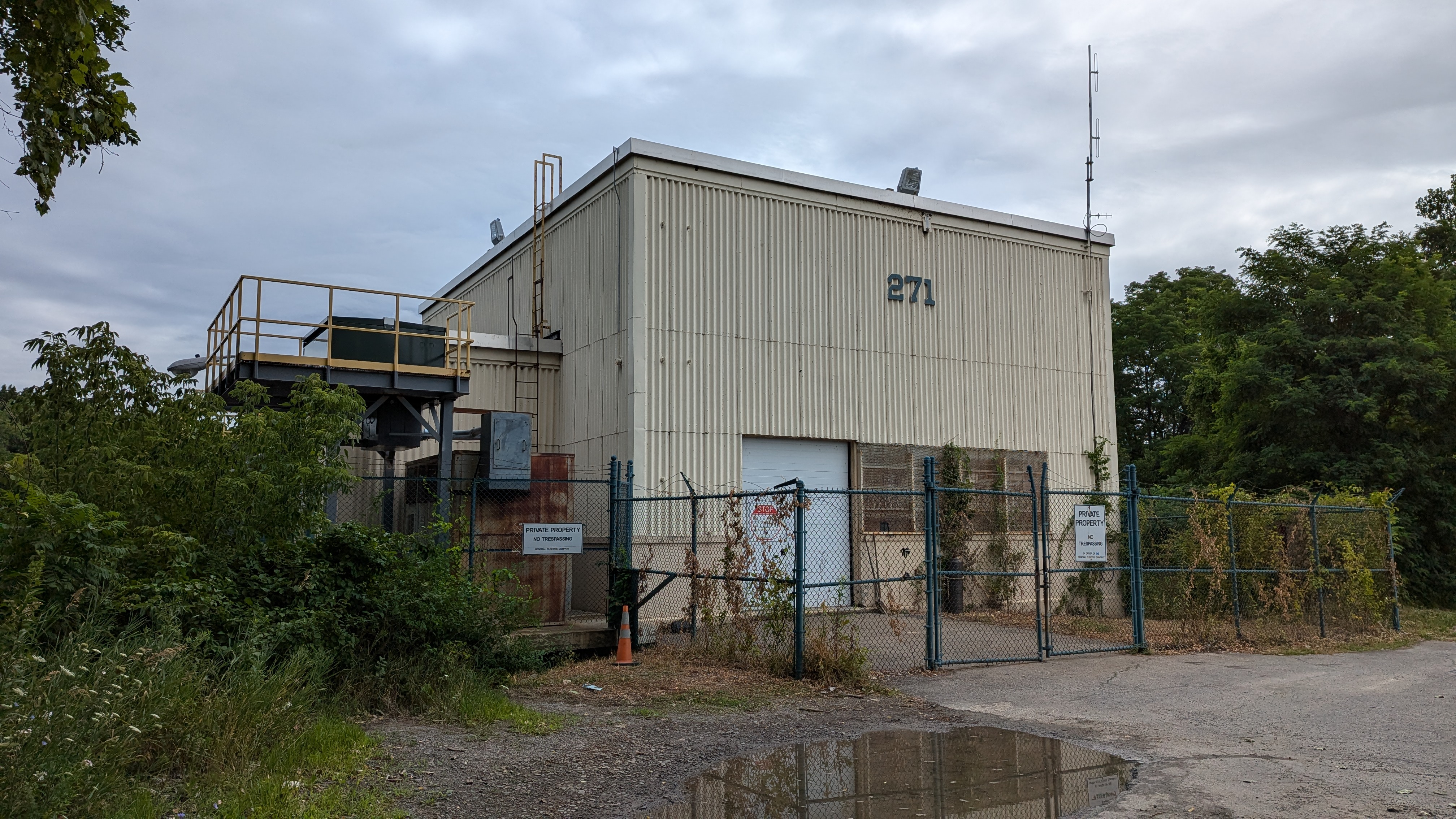 Photo of a yellow metalic building that has a fence going around it. The number 271 is towards the top of the building, and there is barb wiring on the top of the fence