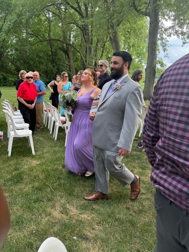 A photo of a man in a grey three piece suit walking down an aisle between rows of people with a woman who is in a purple dress