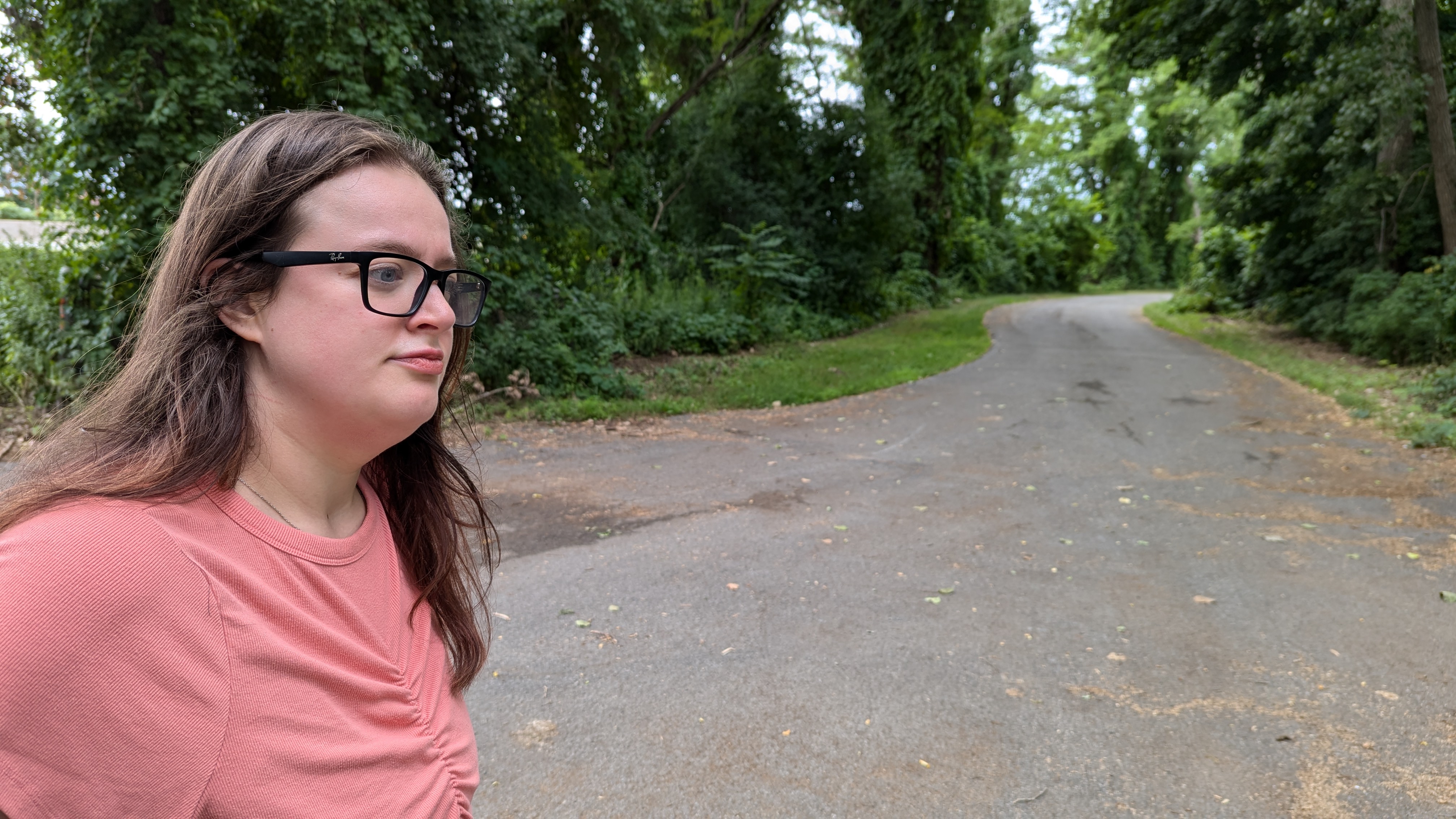 Photo of a paved trail through trees, with a woman in a pink shirt on the left side of the image looking towards the right side of the image