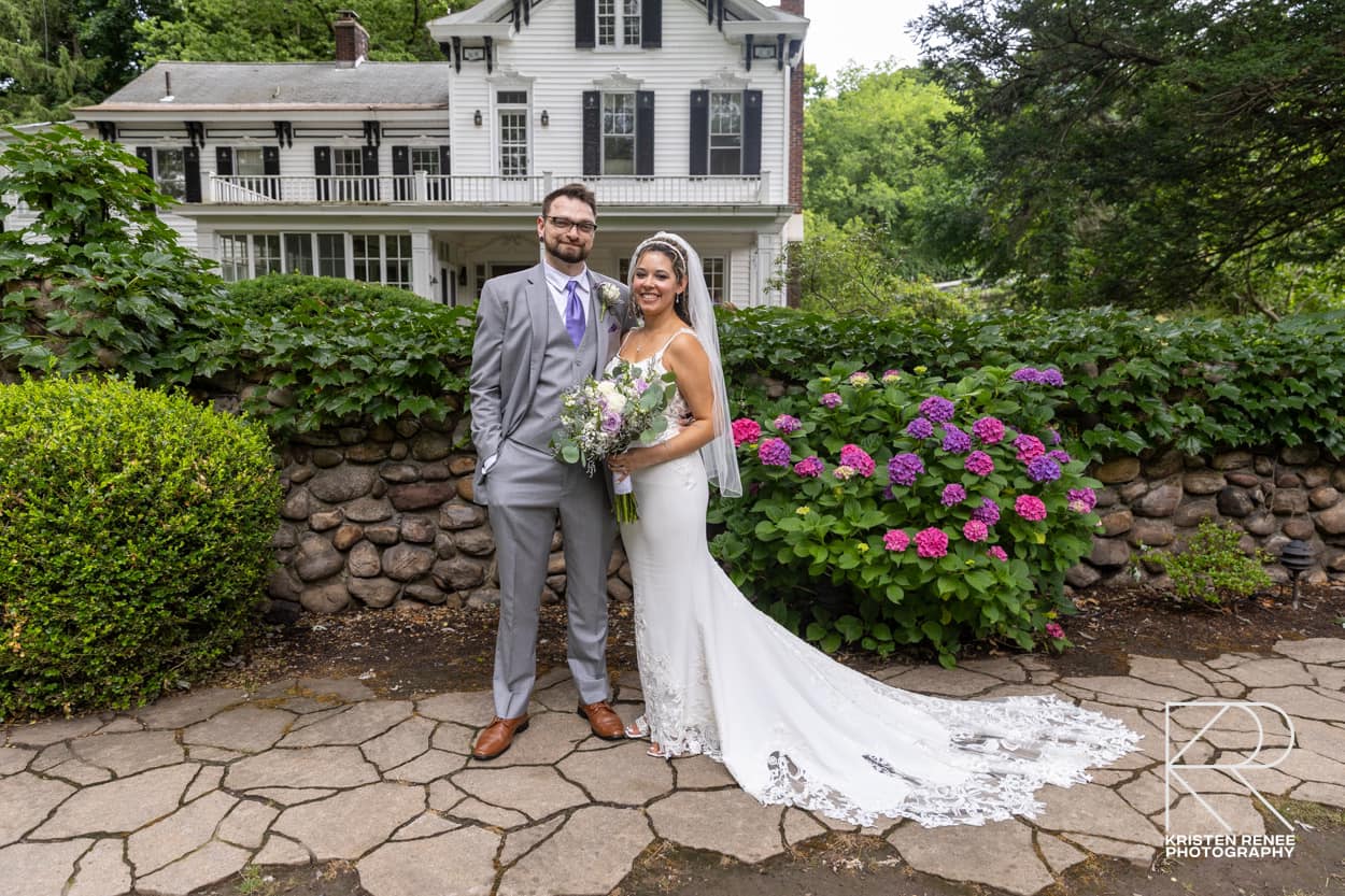 A photo of a bride (Ali) and groom (Tanner) standing in front of an old house. The bride is in a flowing white Dress while the groom is in a grey three piece suit with a purple tie. The bride is holding a bouqet of flowers. There are some flowers blossoming near the bride