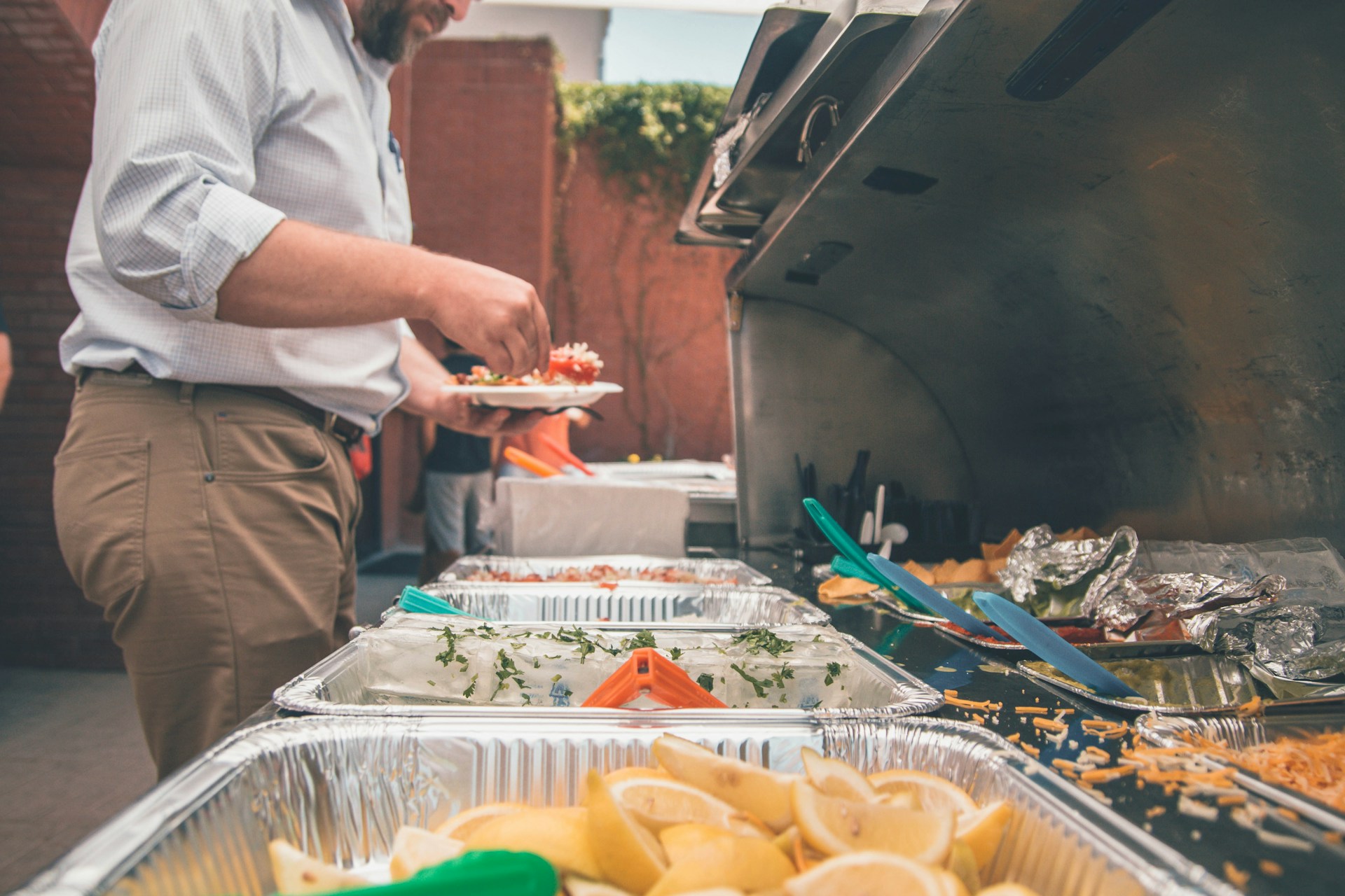 photo of a man at a grill taking some food that is in metal trays and putting it onto his plate