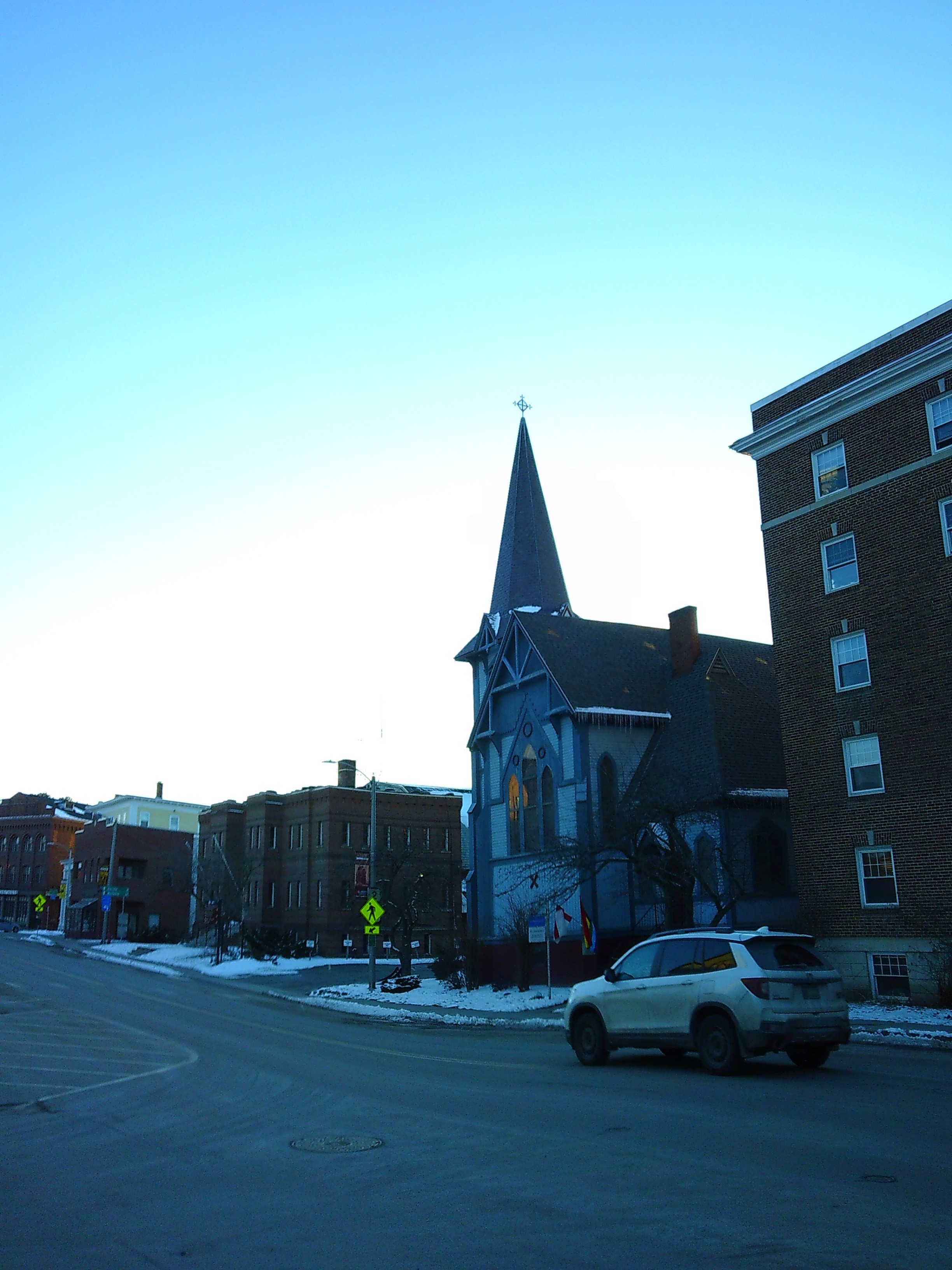 Outside shot of a church with pointed steeple around dusk. It's across the street from where the shot is taken, with brick buildings on either side of it