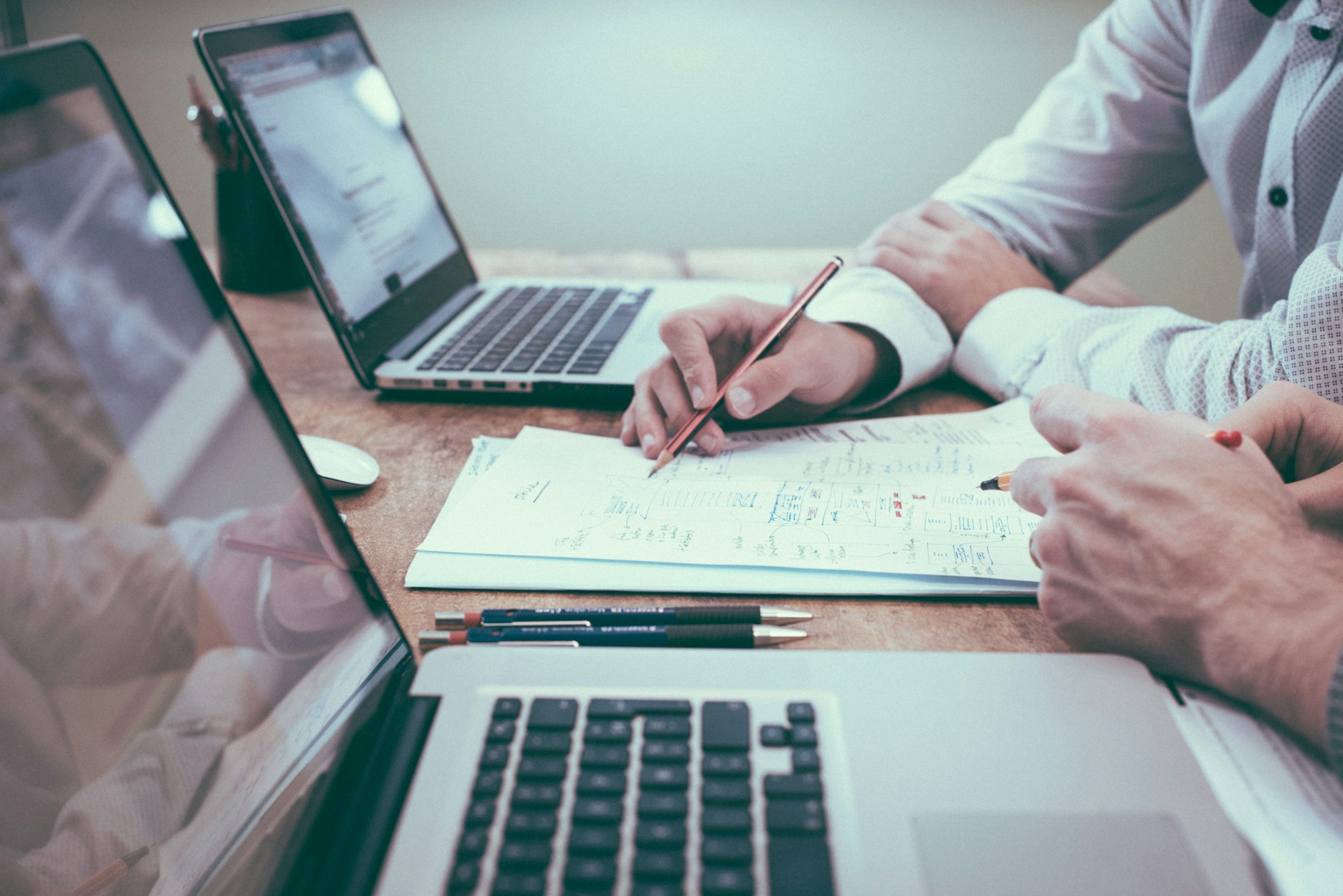 photo of two people at a desk with two laptops in front of them. You can only see from their neck down to about where their bellybuttons would be. They look to be in the middle of a conversation, and there's a notepad between them with writing on it.