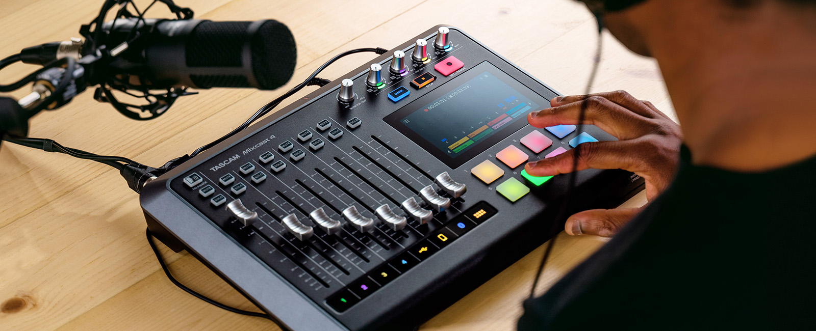 image of person using the tascam mixcast 4. The Tascam Mixcast 4 has 8 audio sliders for the 8 different inputs it can have. It also features a digital screen, as well as 8 colored buttons. There are also knobs along the top right for headphone volume.